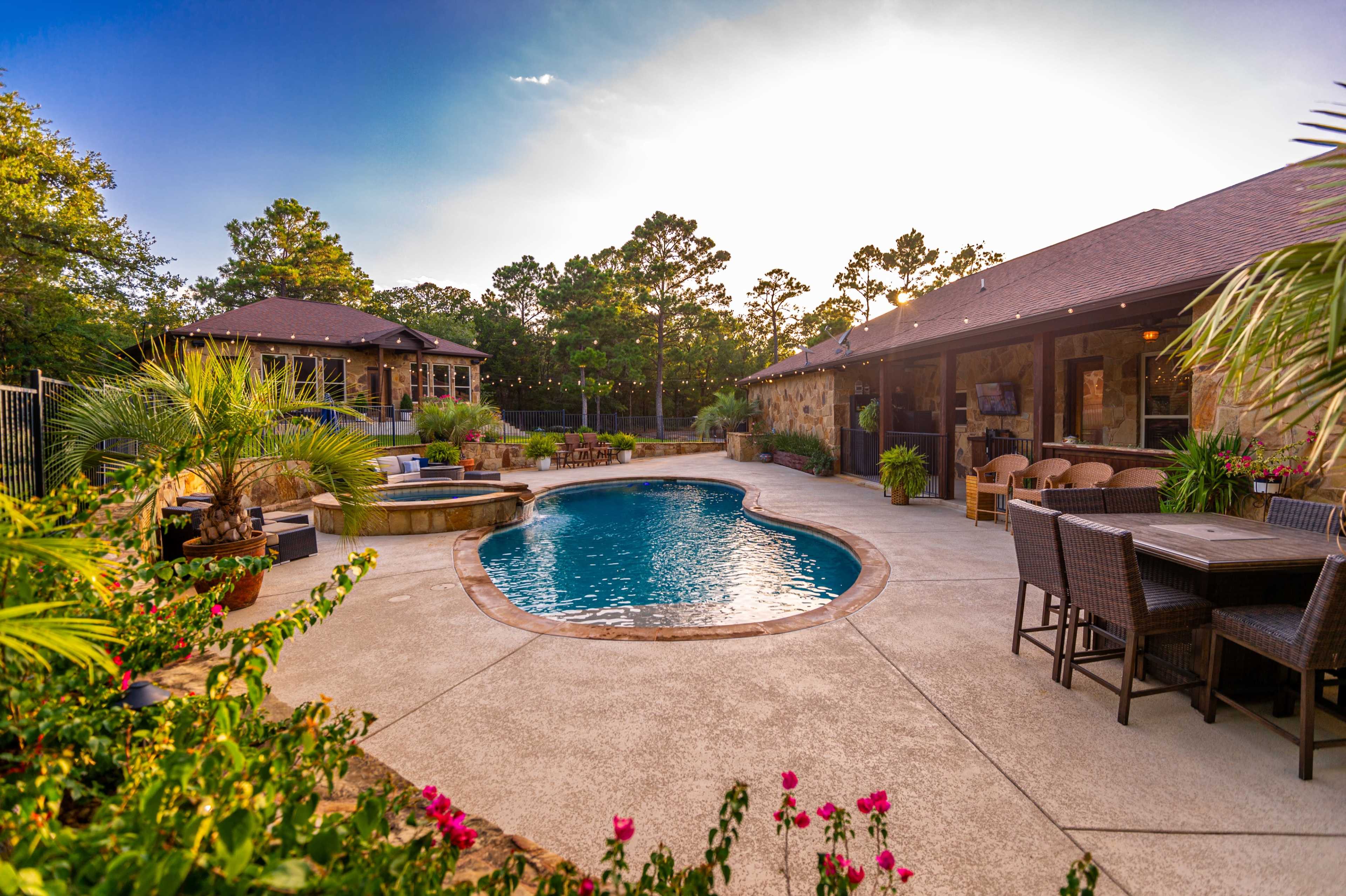 A landscaped backyard features a kidney-shaped swimming pool surrounded by seating areas and a hot tub, with lush greenery and flowers.