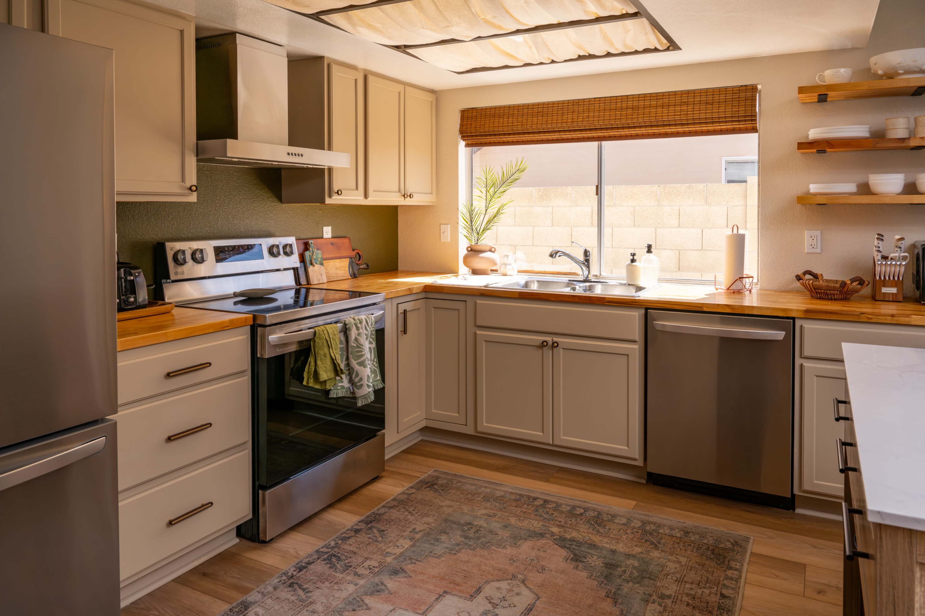 The kitchen features modern appliances, wooden countertops, and natural lighting streaming through a window with a bamboo shade.