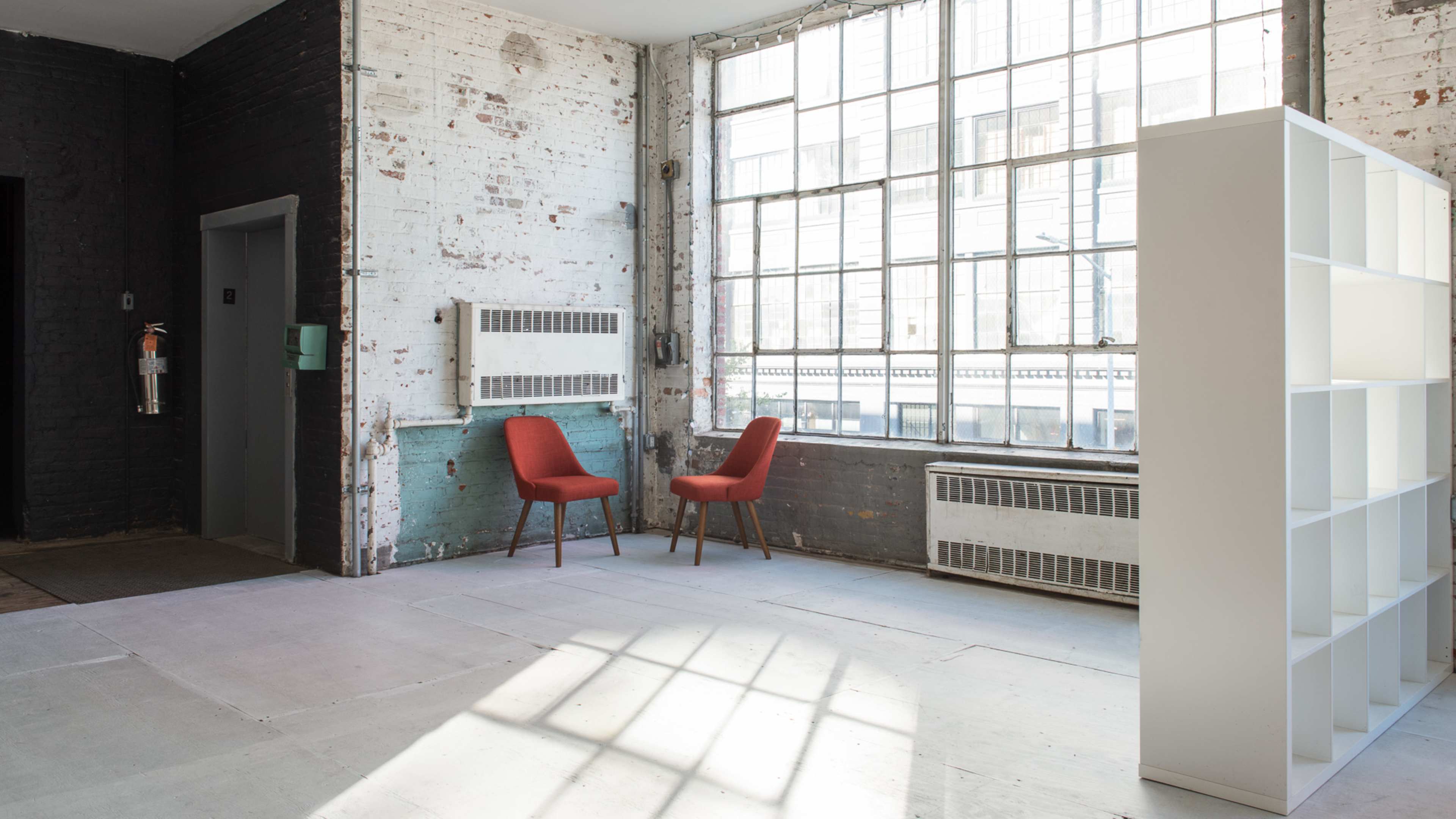 A spacious, minimally furnished room with two red chairs facing a large window and a white shelving unit positioned nearby.