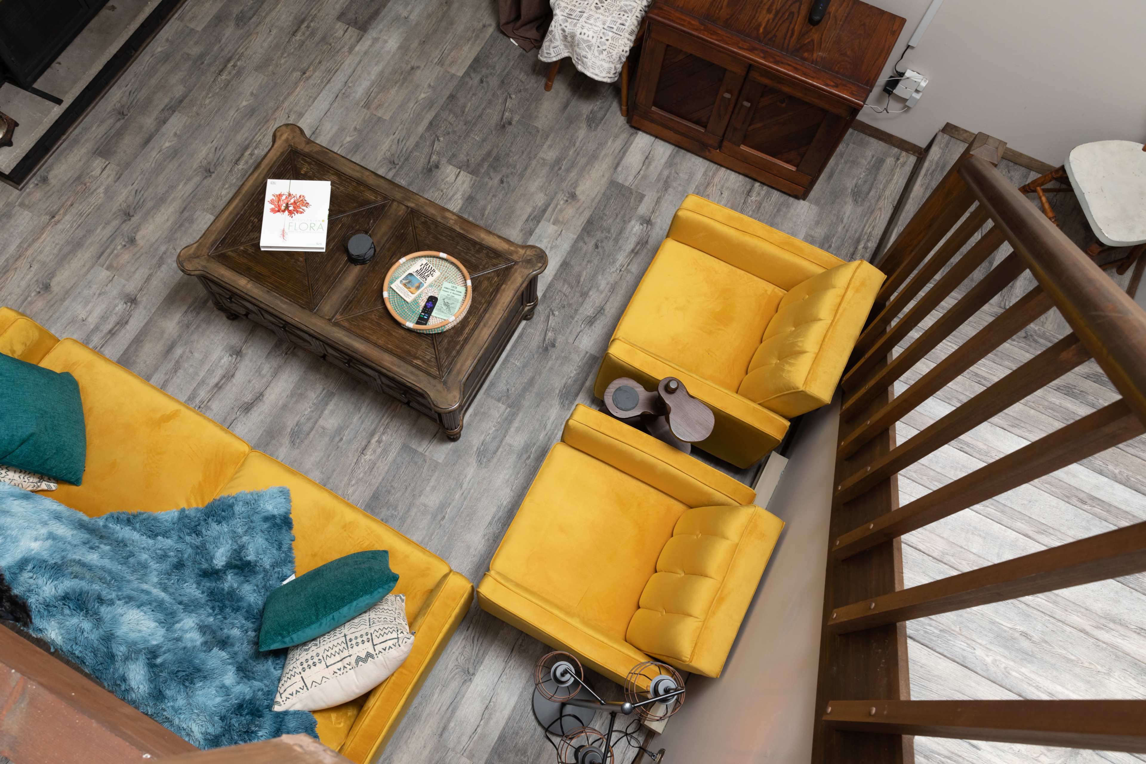 The image shows a cozy living room with yellow armchairs, a wooden coffee table, a turquoise throw, and a wooden railing above.