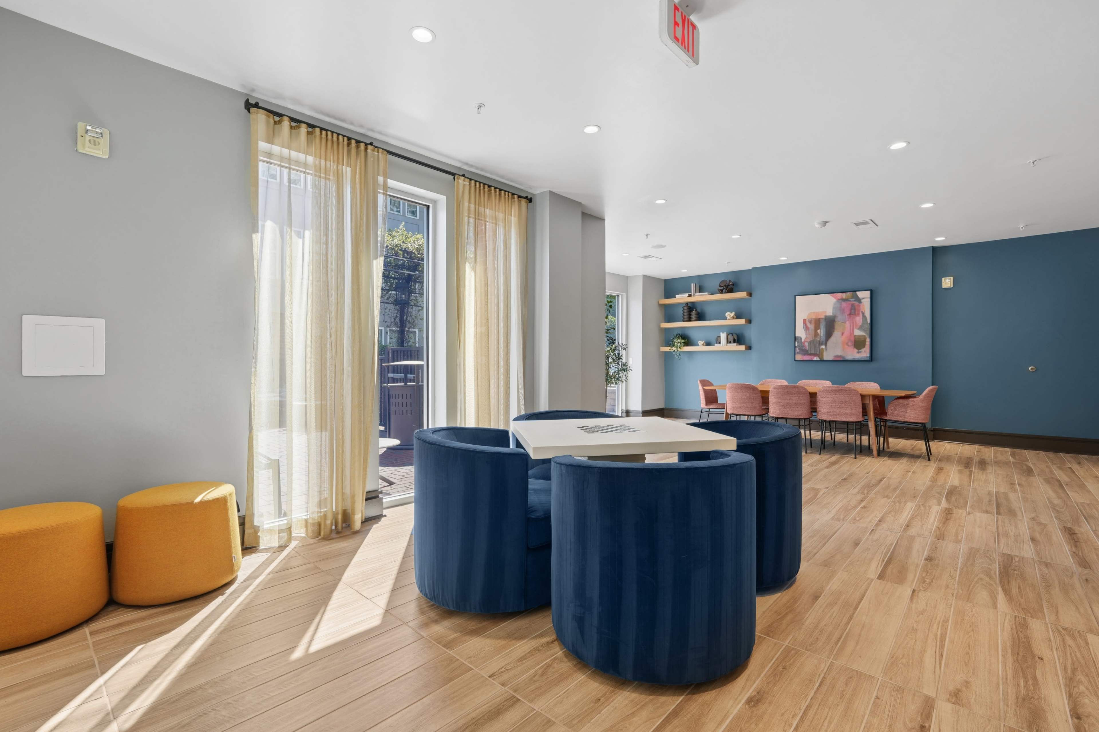 The image shows a modern lounge area with round tables and seating, featuring large windows adorned with curtains, and a dining space with pink chairs against a blue wall.