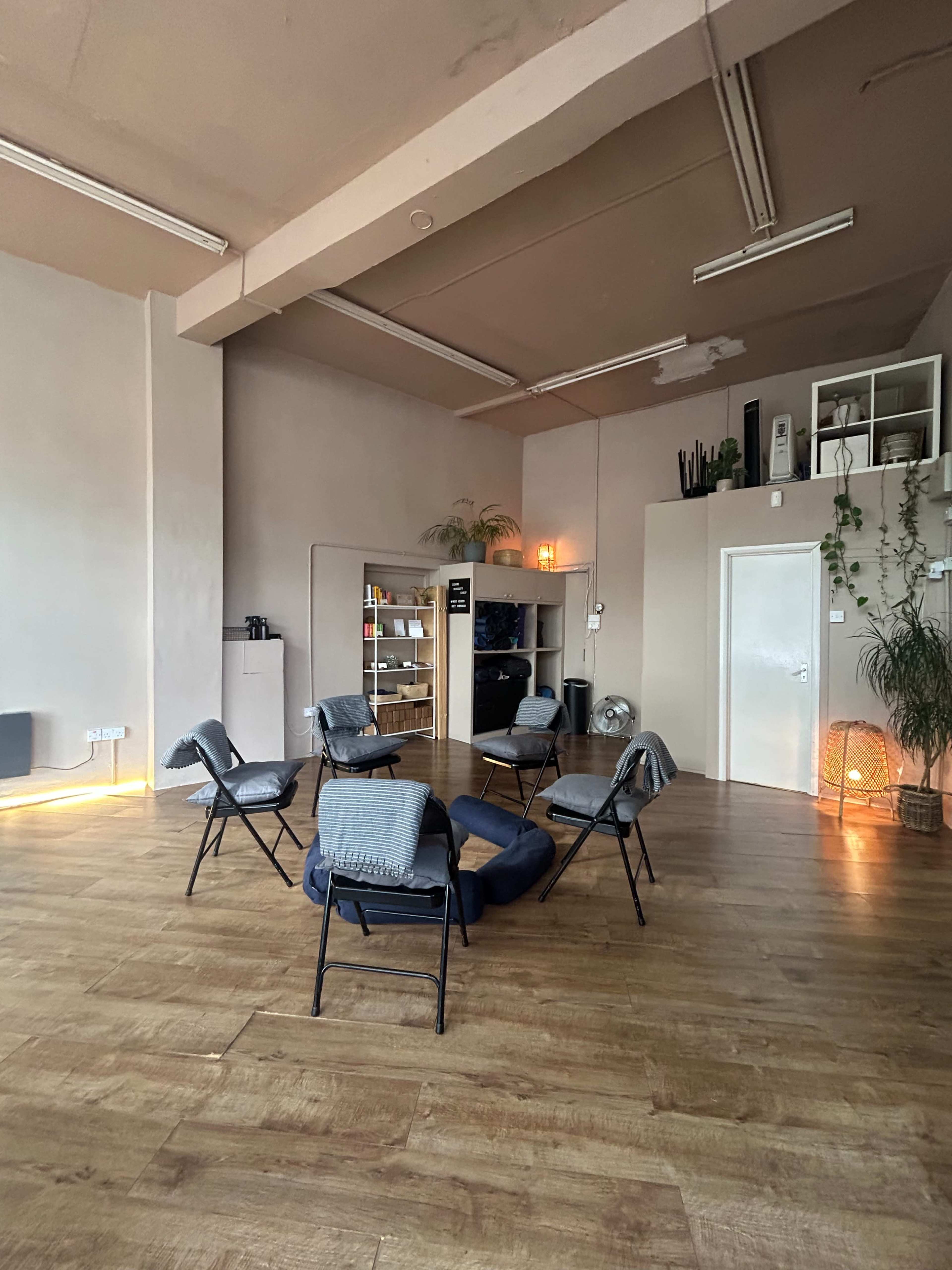 The image shows a spacious room with wooden flooring, featuring a circular arrangement of eight folding chairs, a small cushion in the center, and shelving along the back wall.