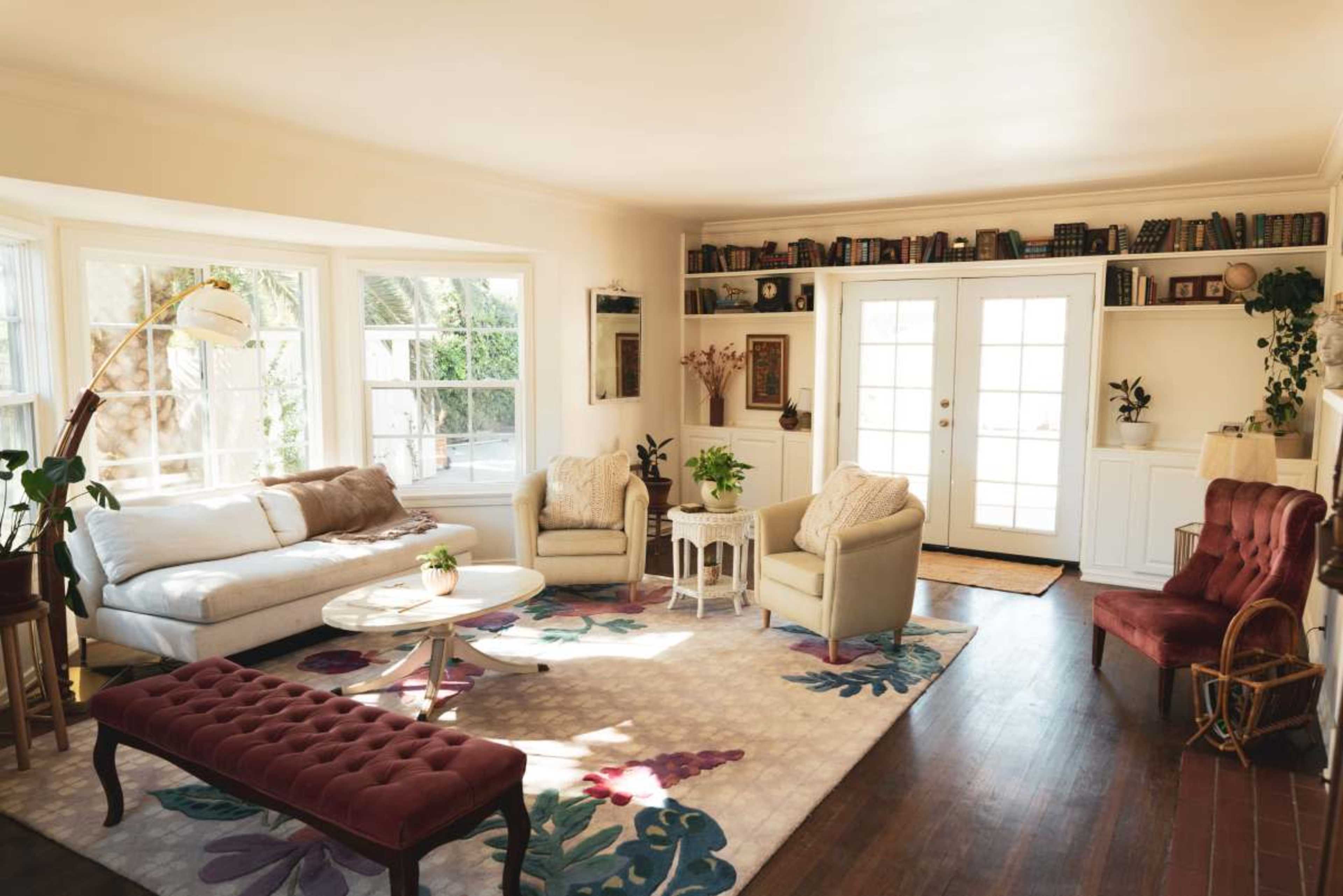 The image shows a bright living room with large windows, a white sofa, two armchairs, and a coffee table on a decorative rug.