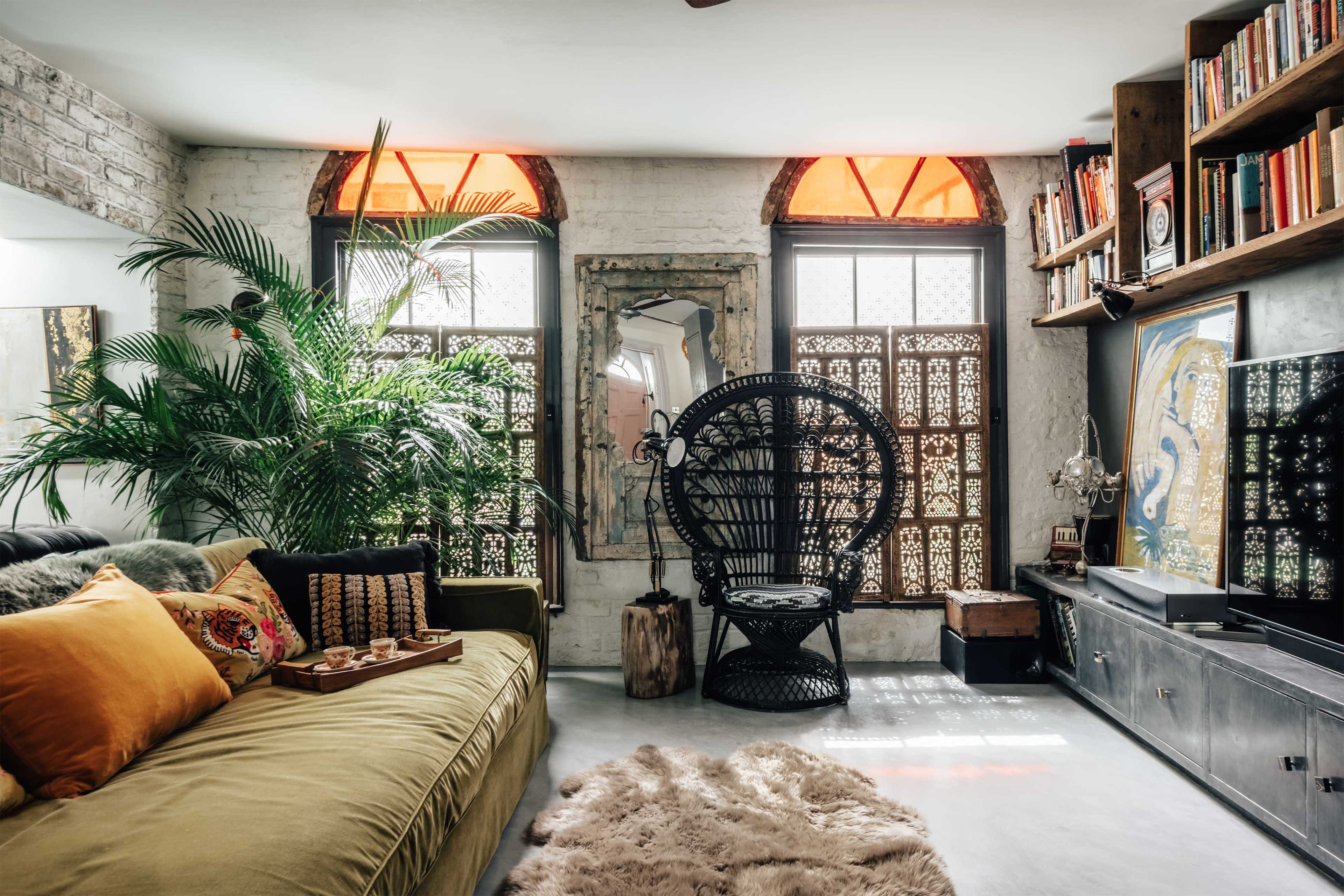A cozy living room features a green sofa adorned with pillows, a large potted plant, and an ornate wicker chair, with bright windows illuminating the space.