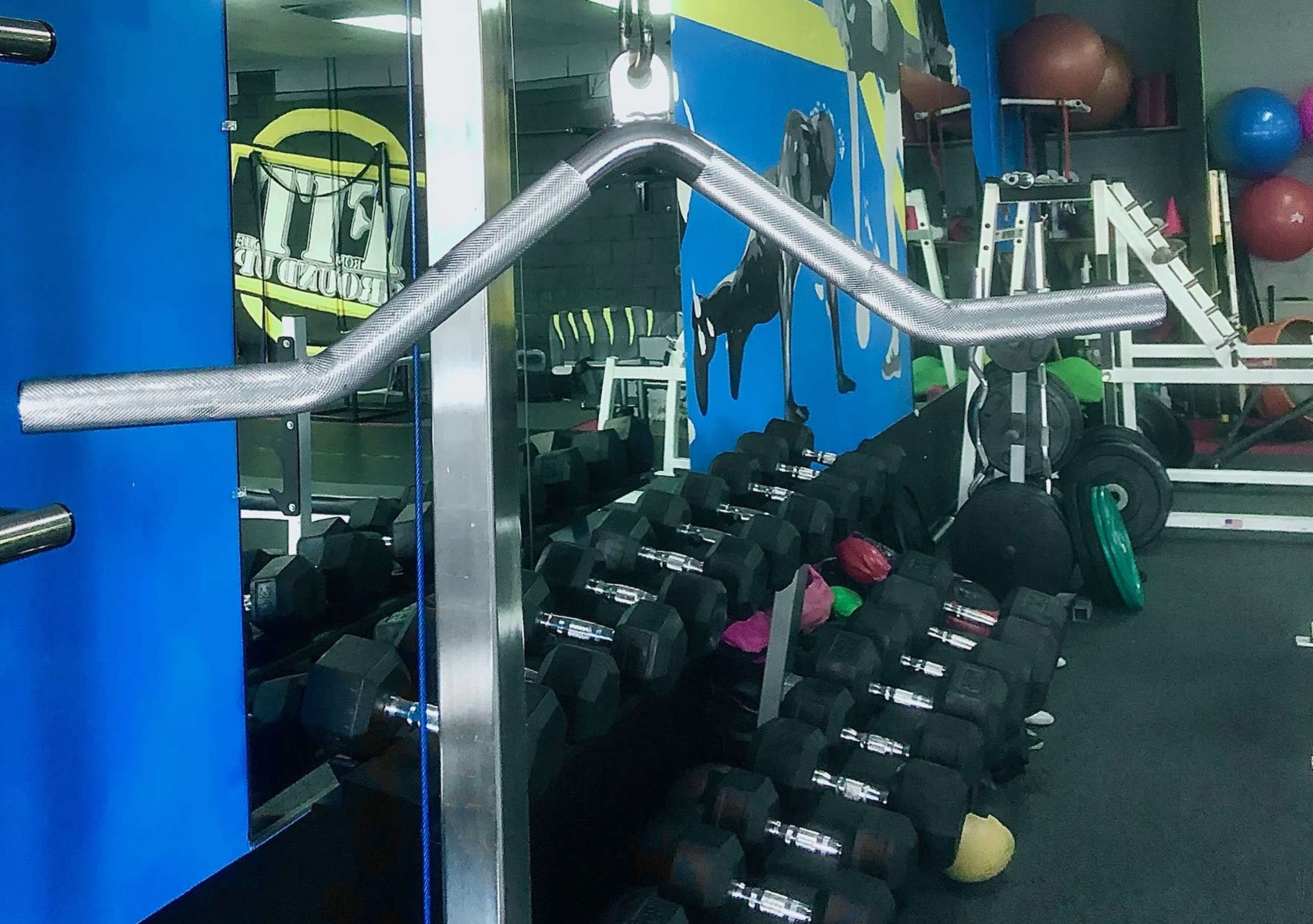 A close-up of a barbell with a unique grip design in a gym, showcasing a rack of dumbbells against a colorful wall.