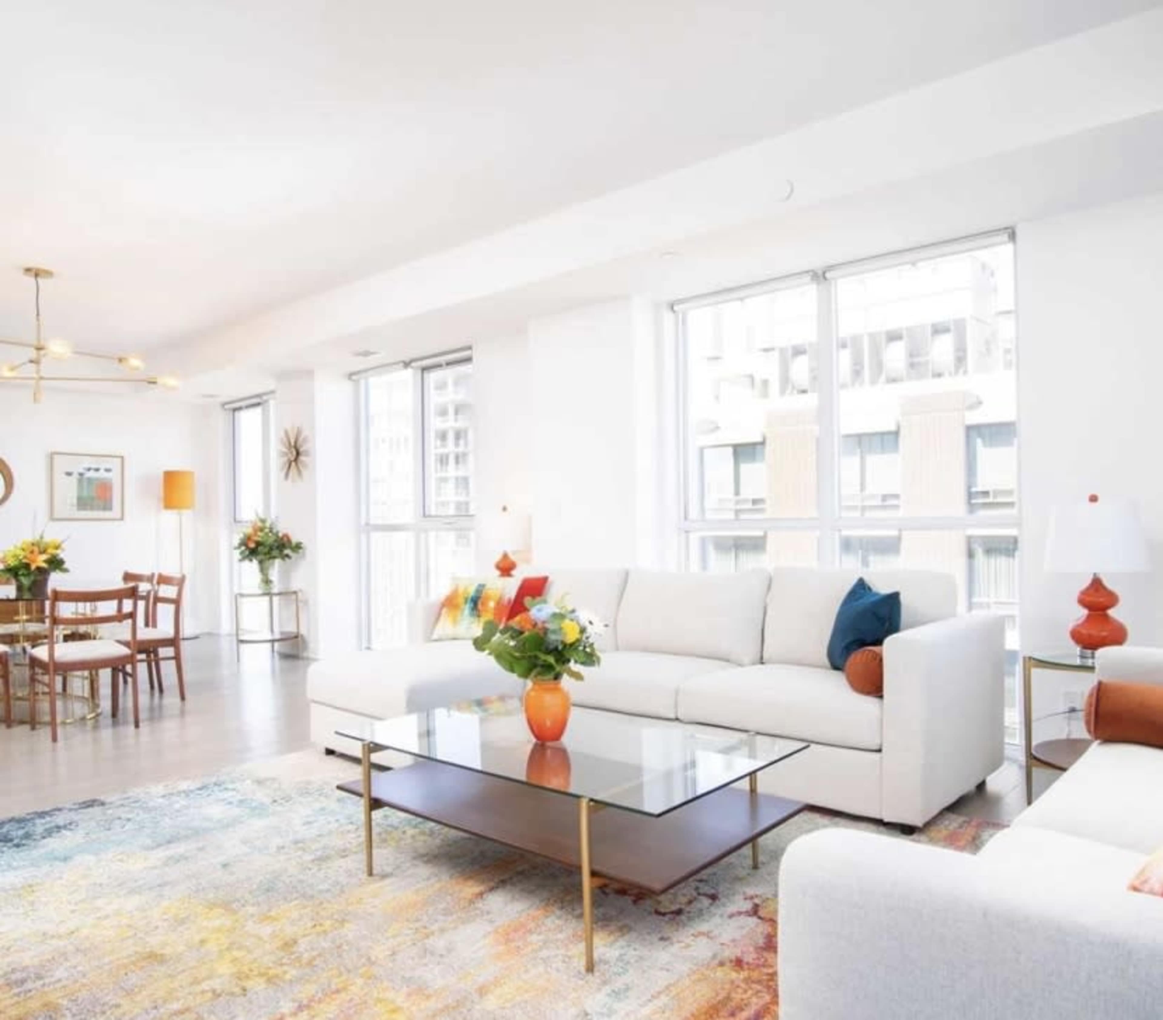 A bright living room features a white sofa, a glass coffee table, and large windows with a view of an outdoor cityscape.