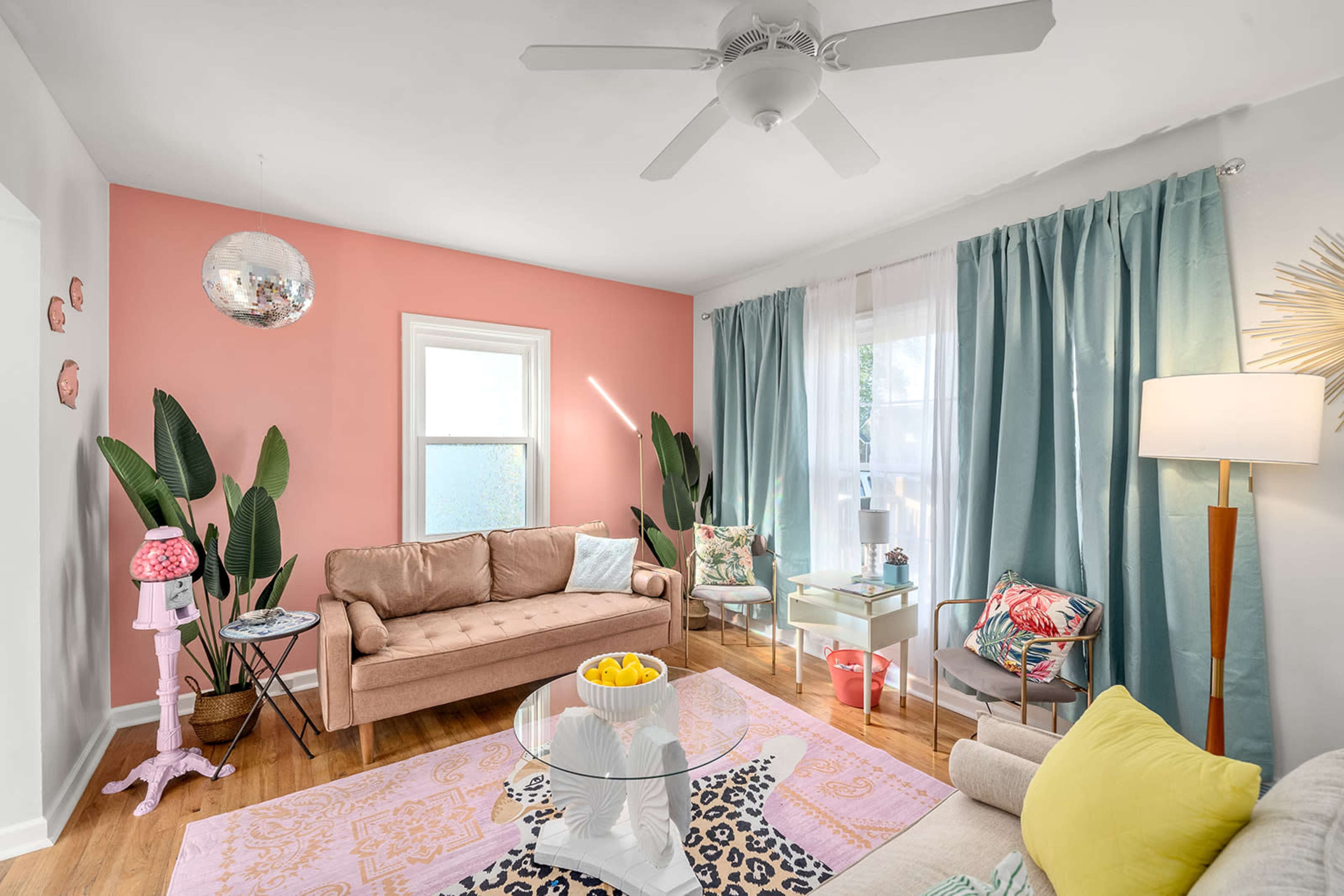 The living room features a pink accent wall, light-colored sofas, potted plants, and a large rug with a mix of patterns.