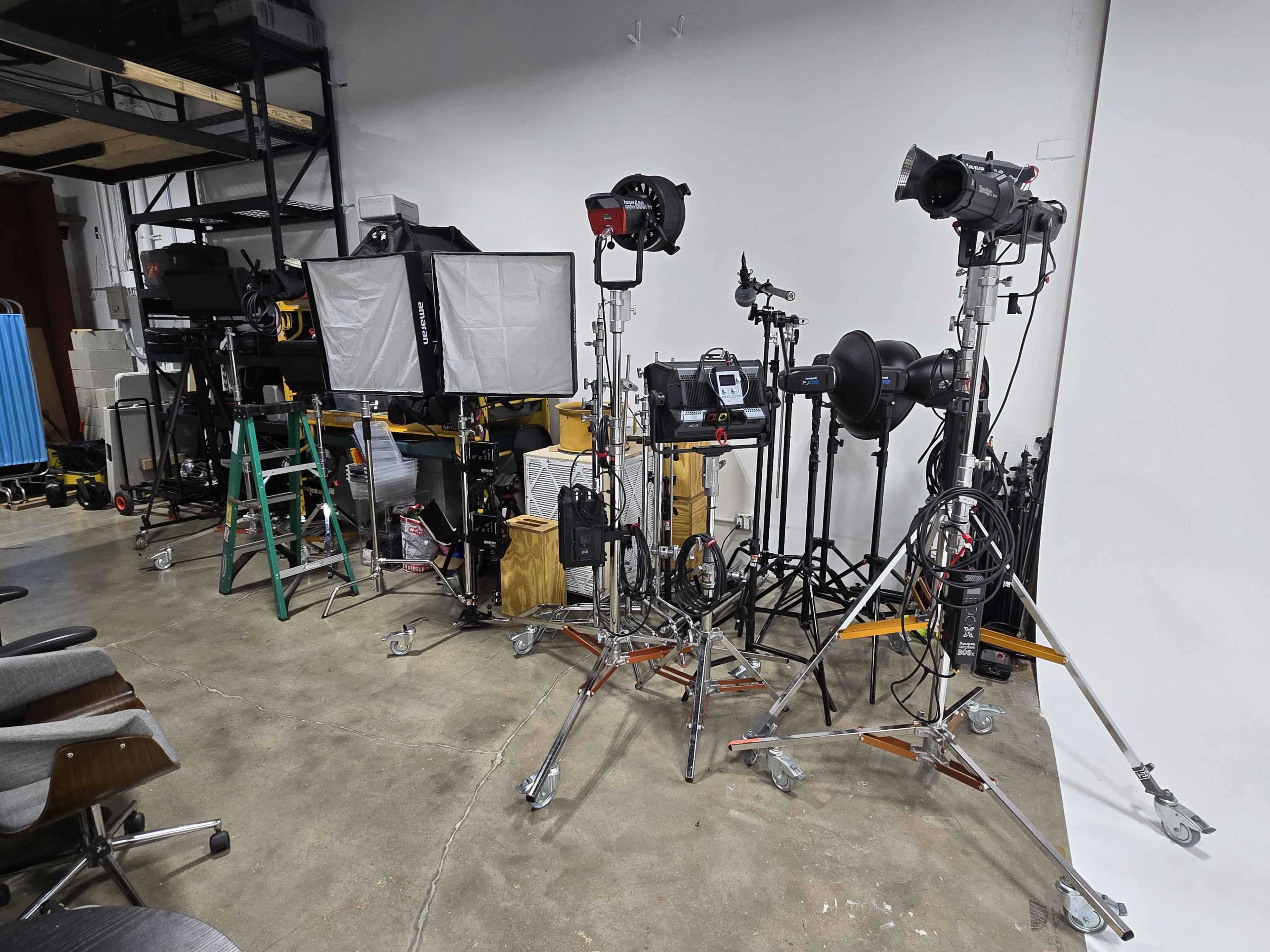 The image shows a collection of photography equipment, including lights, stands, and a ladder, organized in a studio setting with a plain backdrop.