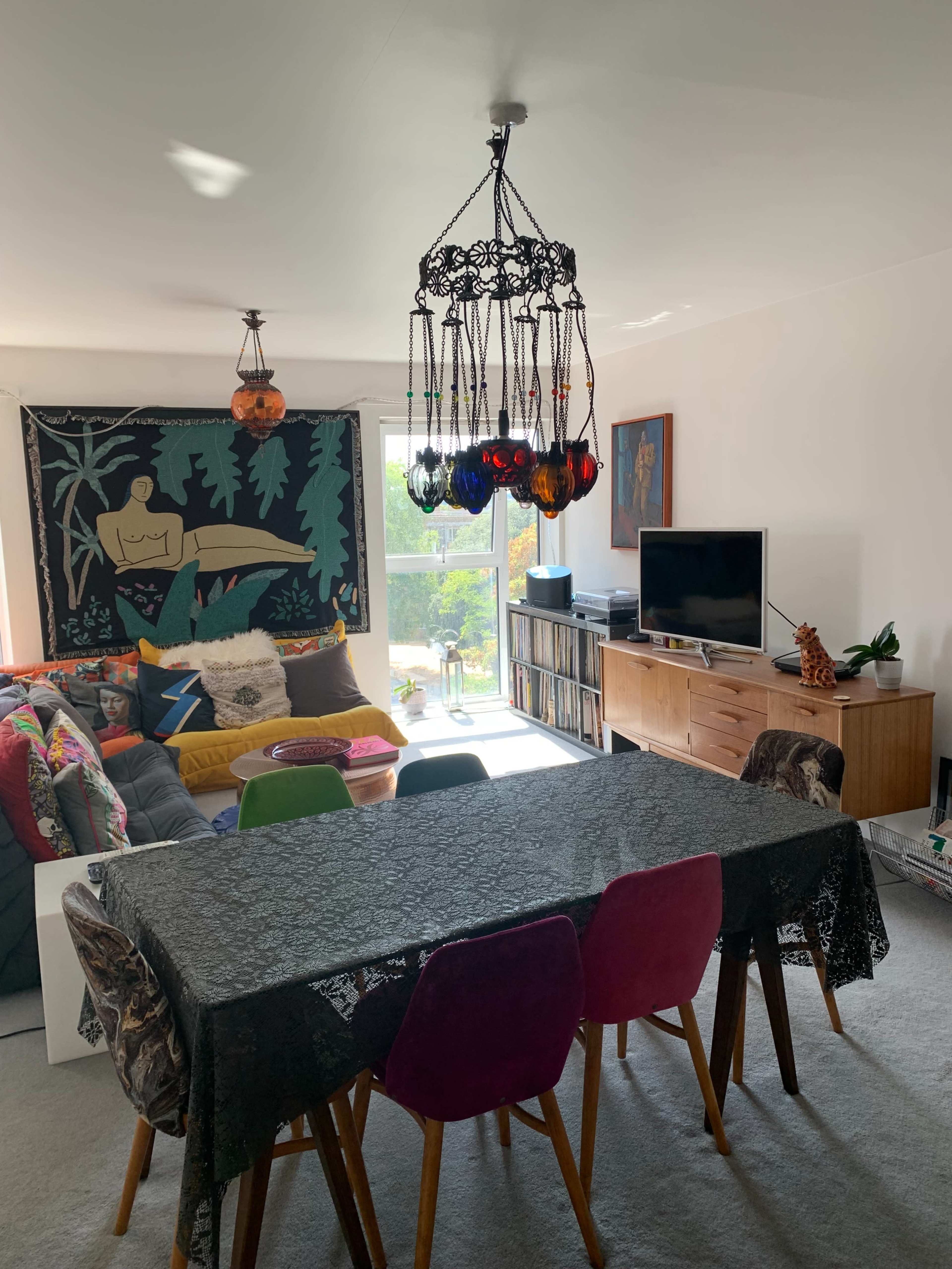 The image shows a living room with a dining table surrounded by colored chairs, a couch with decorative pillows, a wall tapestry, and a television on a wooden cabinet.