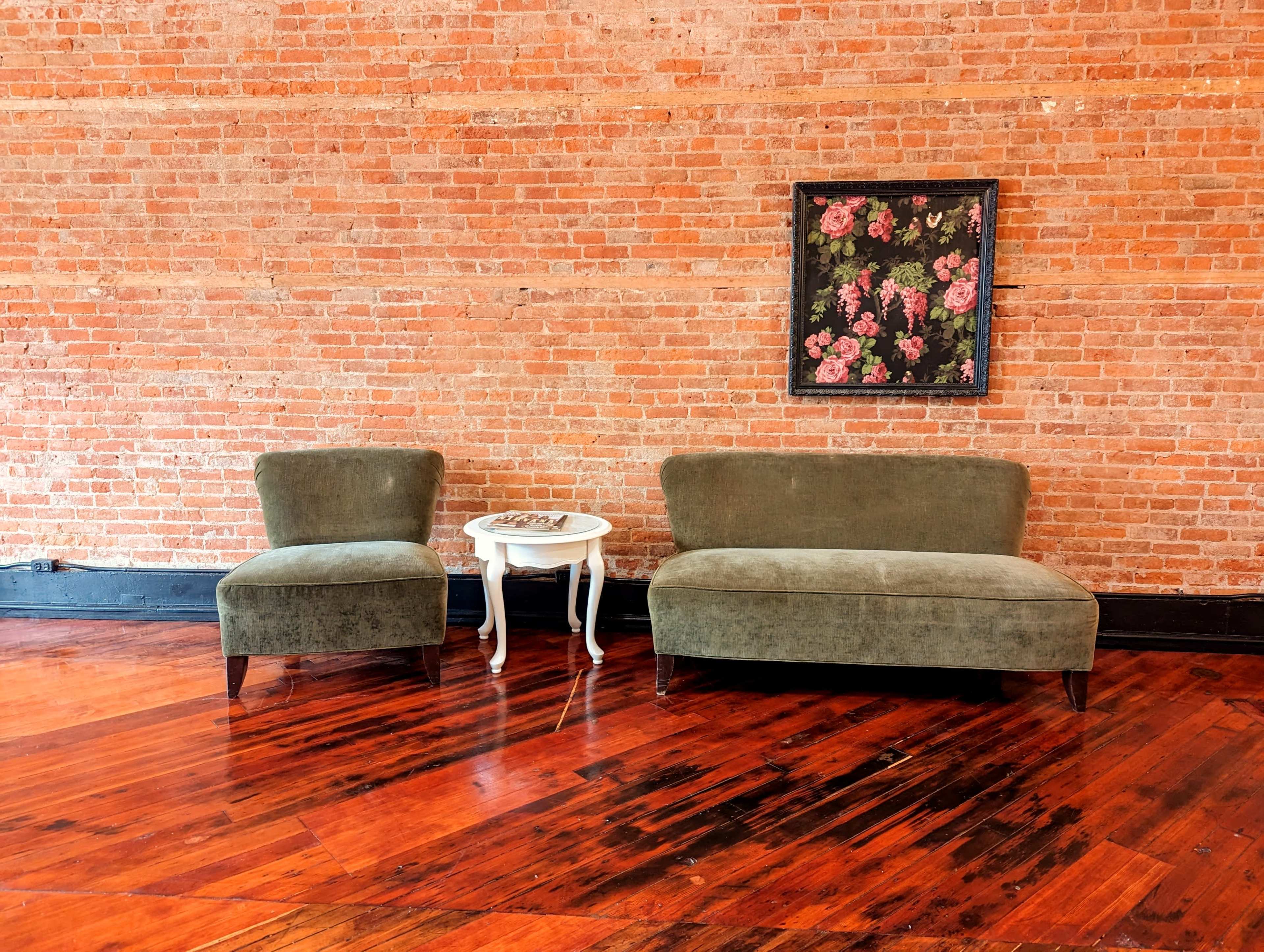 A seating area features two green upholstered chairs and a small white side table against a brick wall, with a floral painting above.