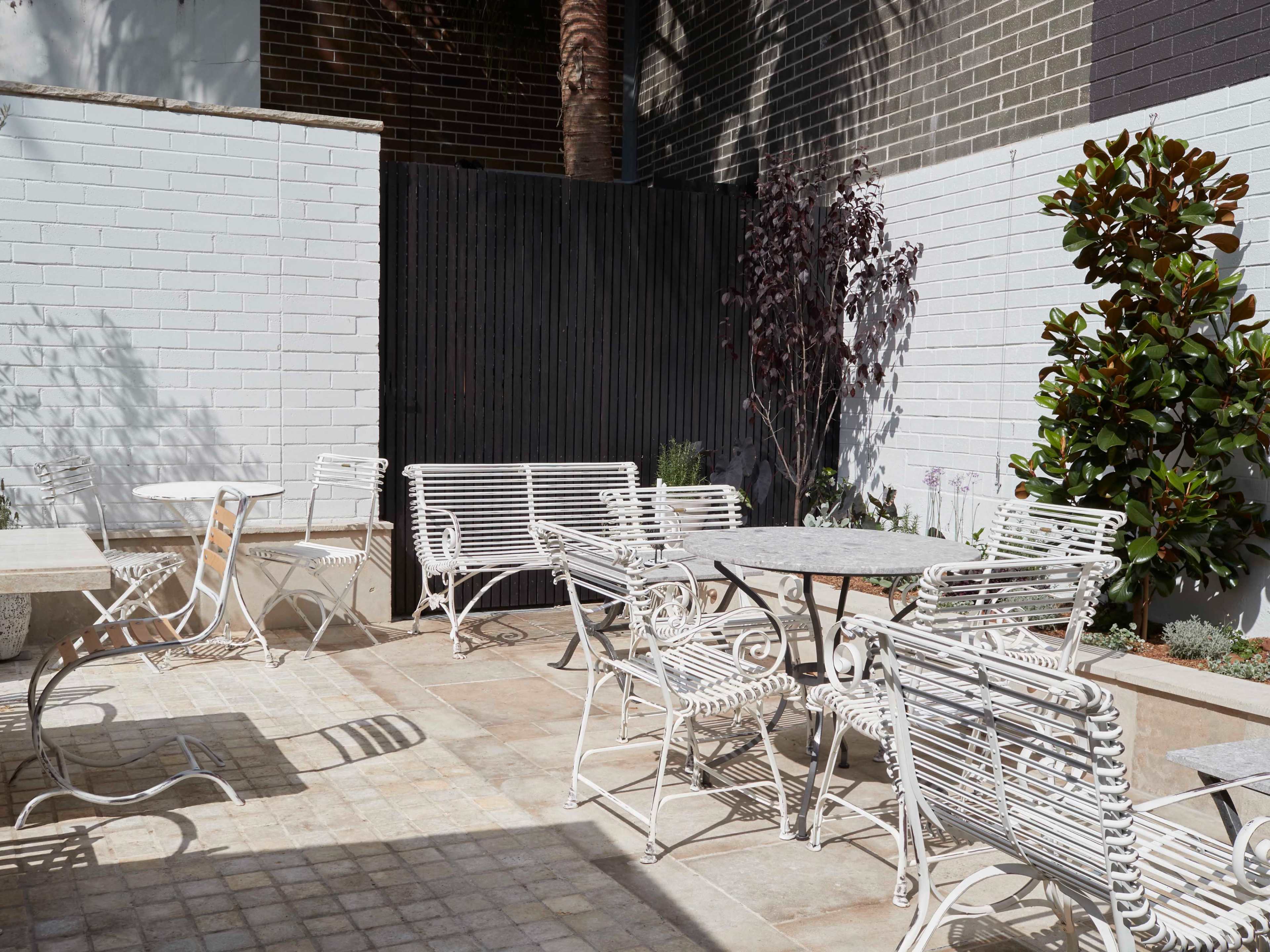 A courtyard features multiple white metal tables and chairs surrounded by greenery and textured walls.