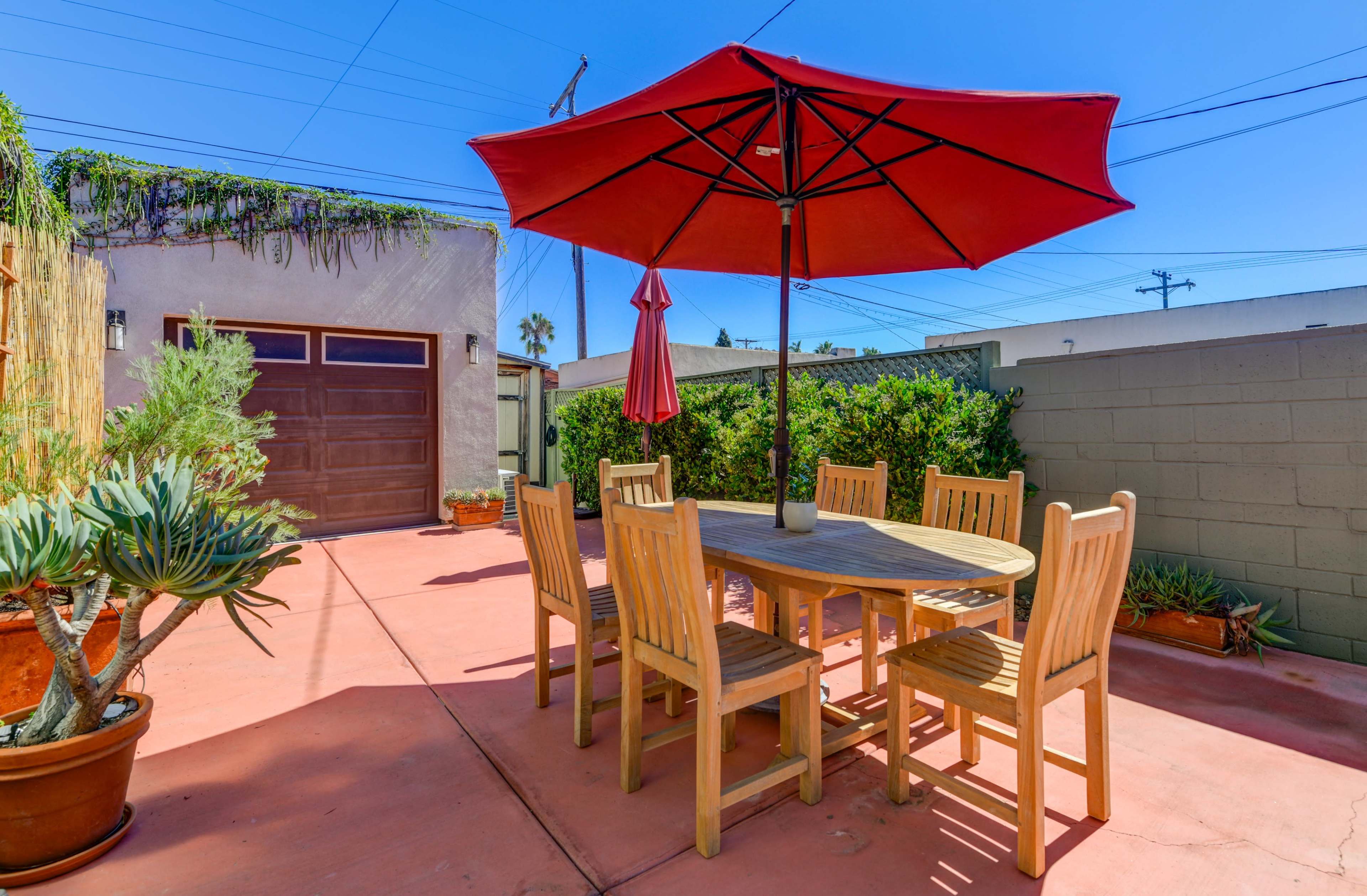 A wooden dining table with six chairs is set under a red umbrella in a patio area surrounded by potted plants and a garage.