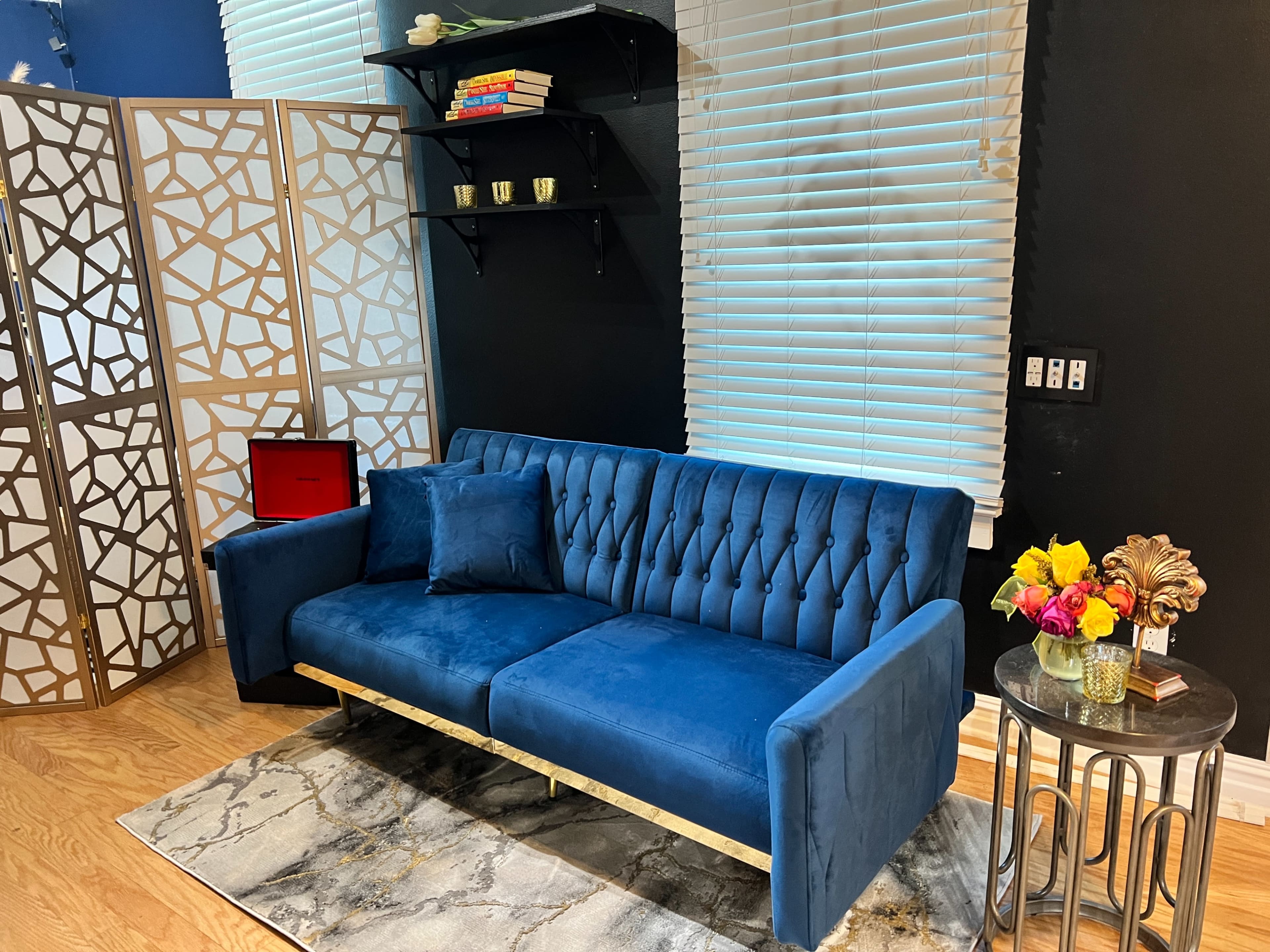 The image shows a blue velvet sofa with tufted upholstery, positioned near a decorative screen, shelves with books, and a side table with flowers.