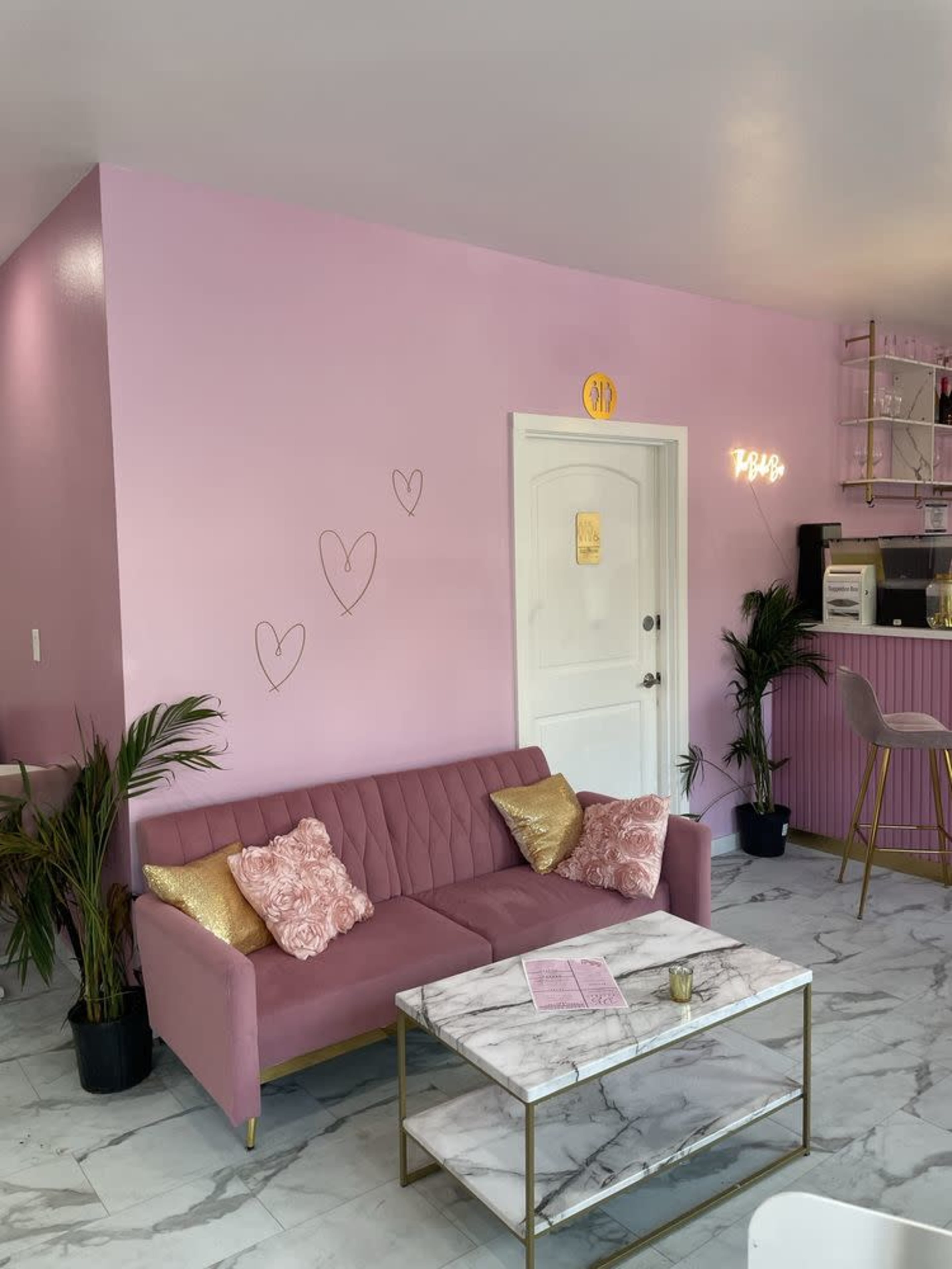 The image shows a cozy interior of a pink-themed cafe featuring a pink couch, a marble coffee table, and a bar counter in the background.