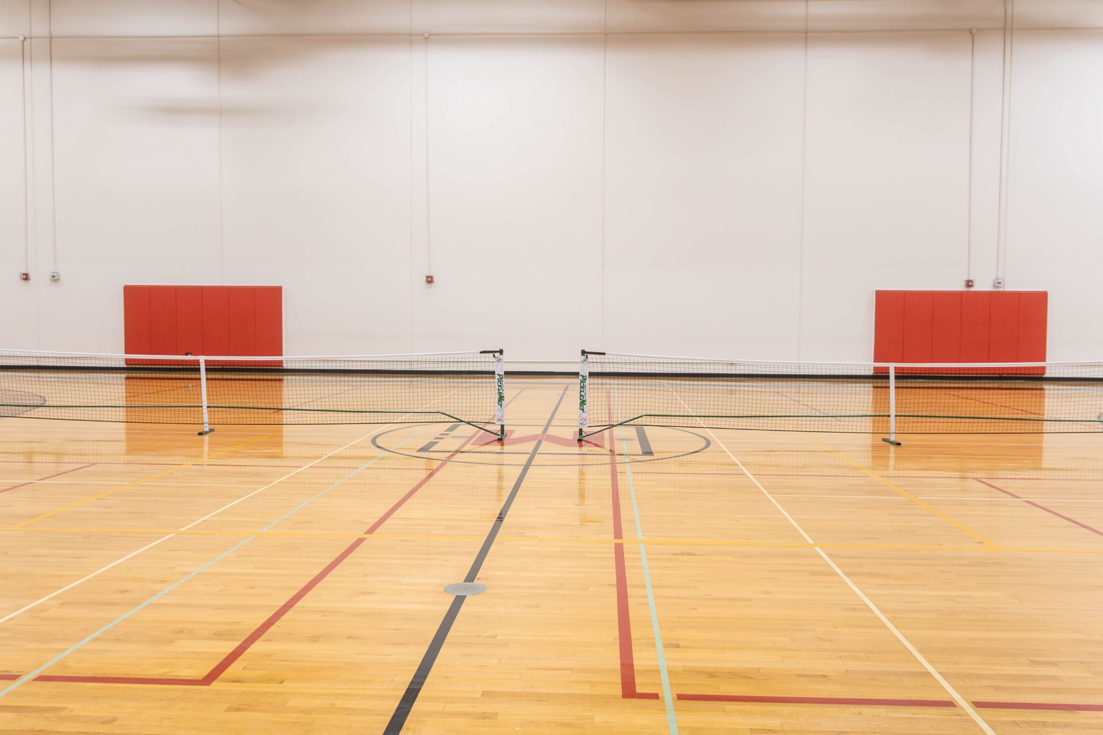The image shows two tennis nets set up on a wooden gymnasium floor with red padding on the walls.