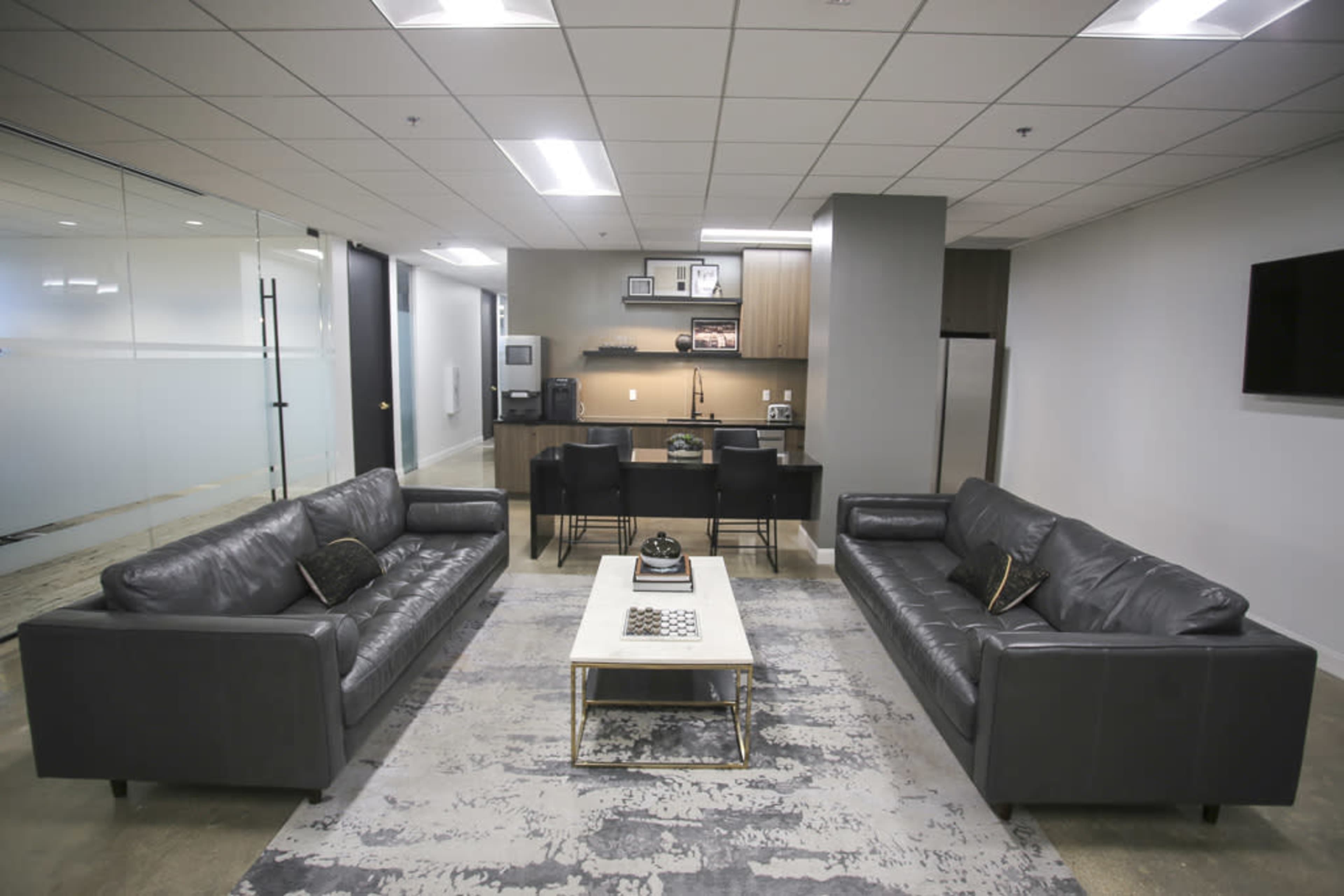 A modern office lounge with two leather sofas facing a central coffee table, surrounded by a rug, with a kitchenette visible in the background.