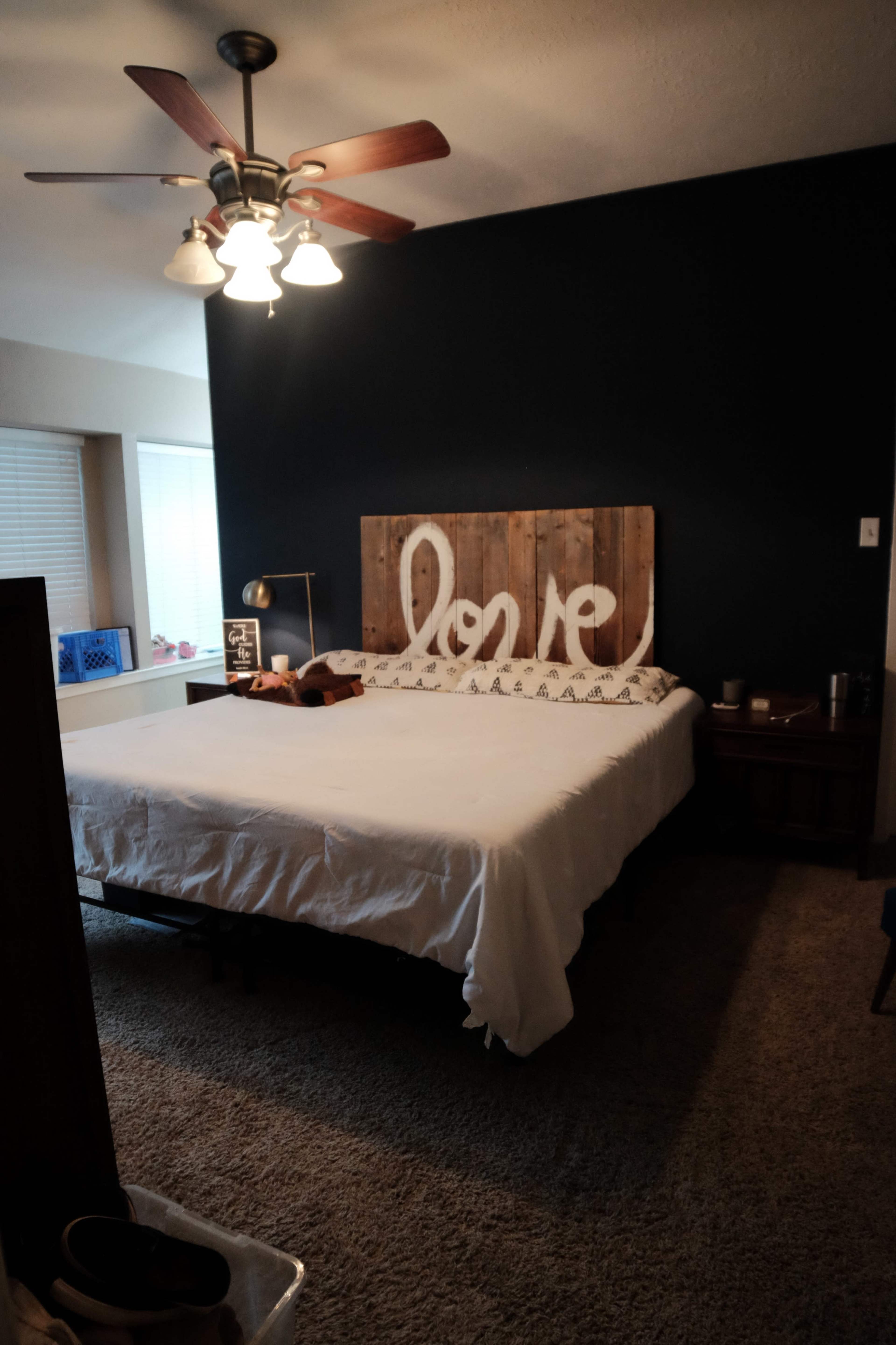 A bedroom with a bed featuring a wooden headboard that has the word "love" painted on it, flanked by a ceiling fan and a dark blue wall.