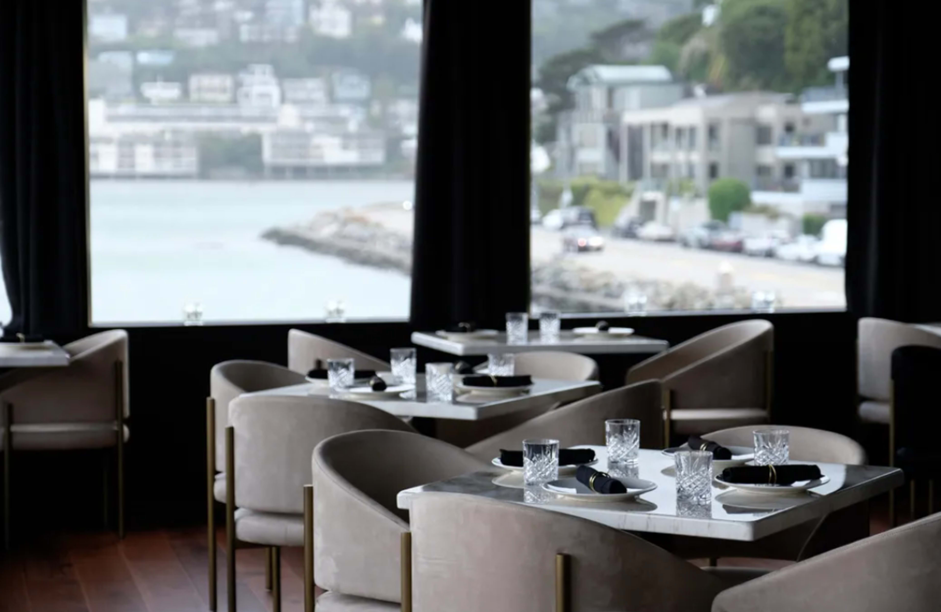 A modern restaurant features elegantly set tables with glassware, overlooking a coastal view through large windows.
