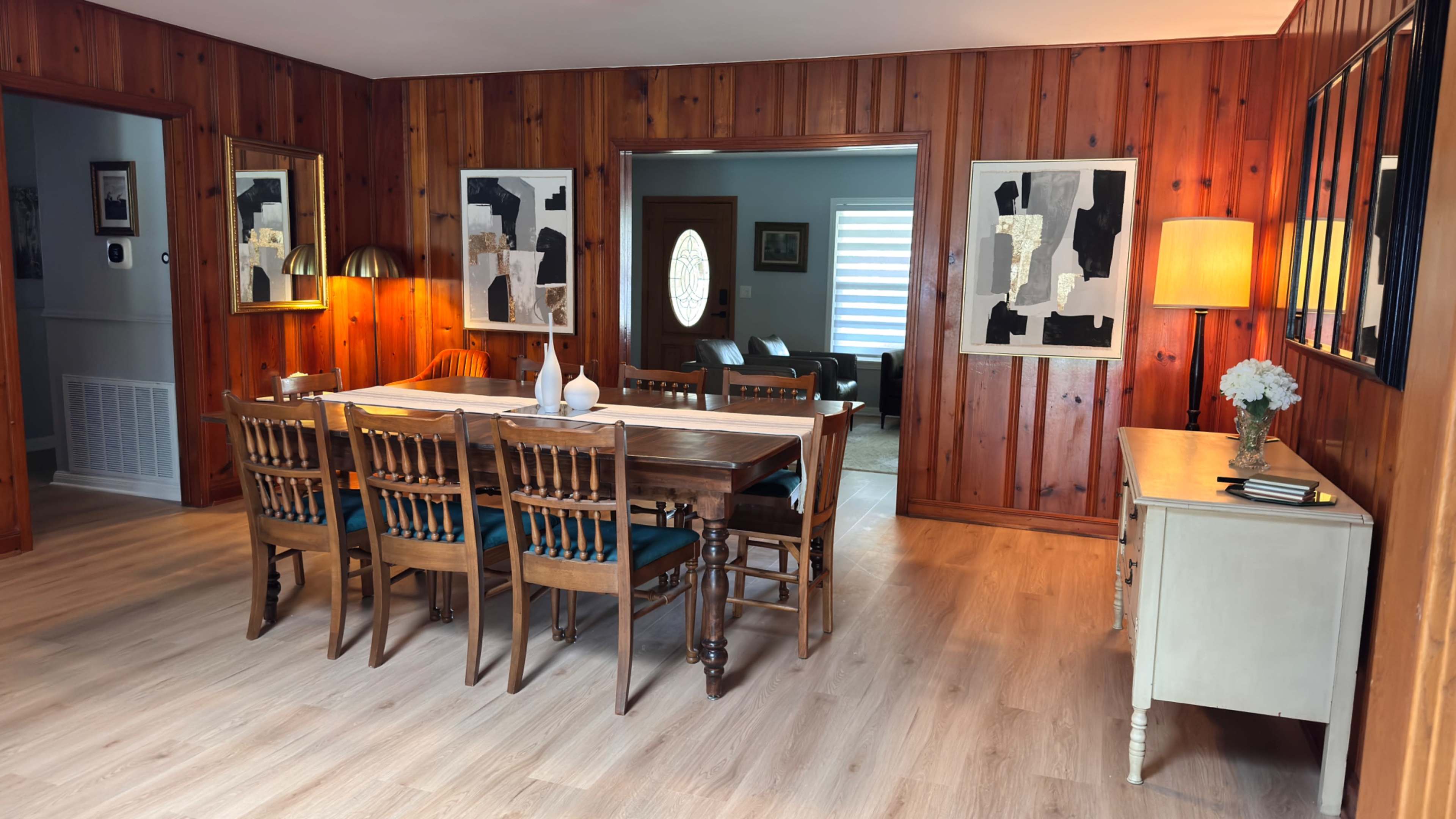 The image shows a cozy dining area with a long wooden table surrounded by chairs, framed by wooden walls, and accented by decorative artwork and lamps.