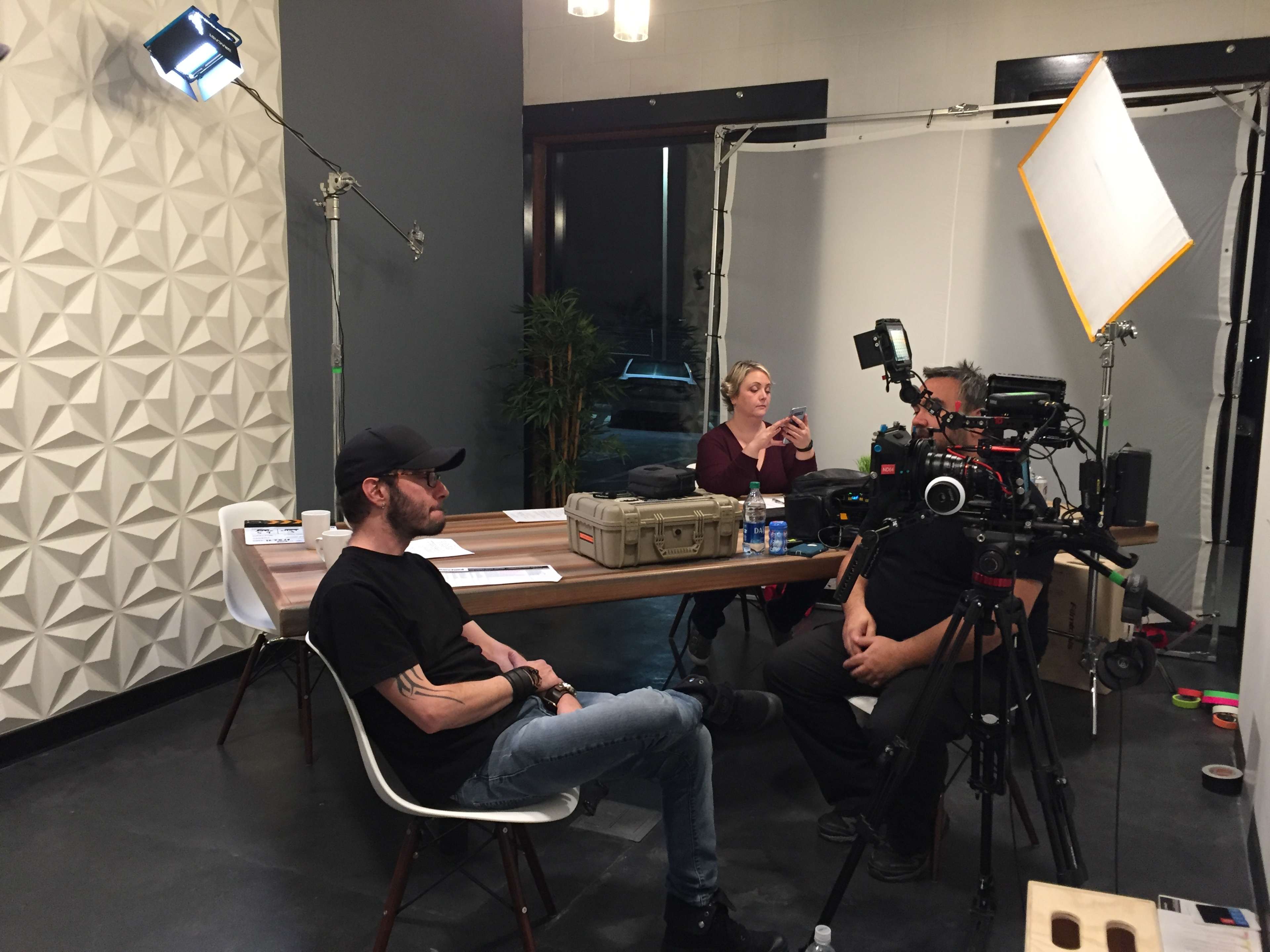 A man sits in a chair near a table while a woman uses her phone and a crew sets up camera equipment in a brightly lit room.