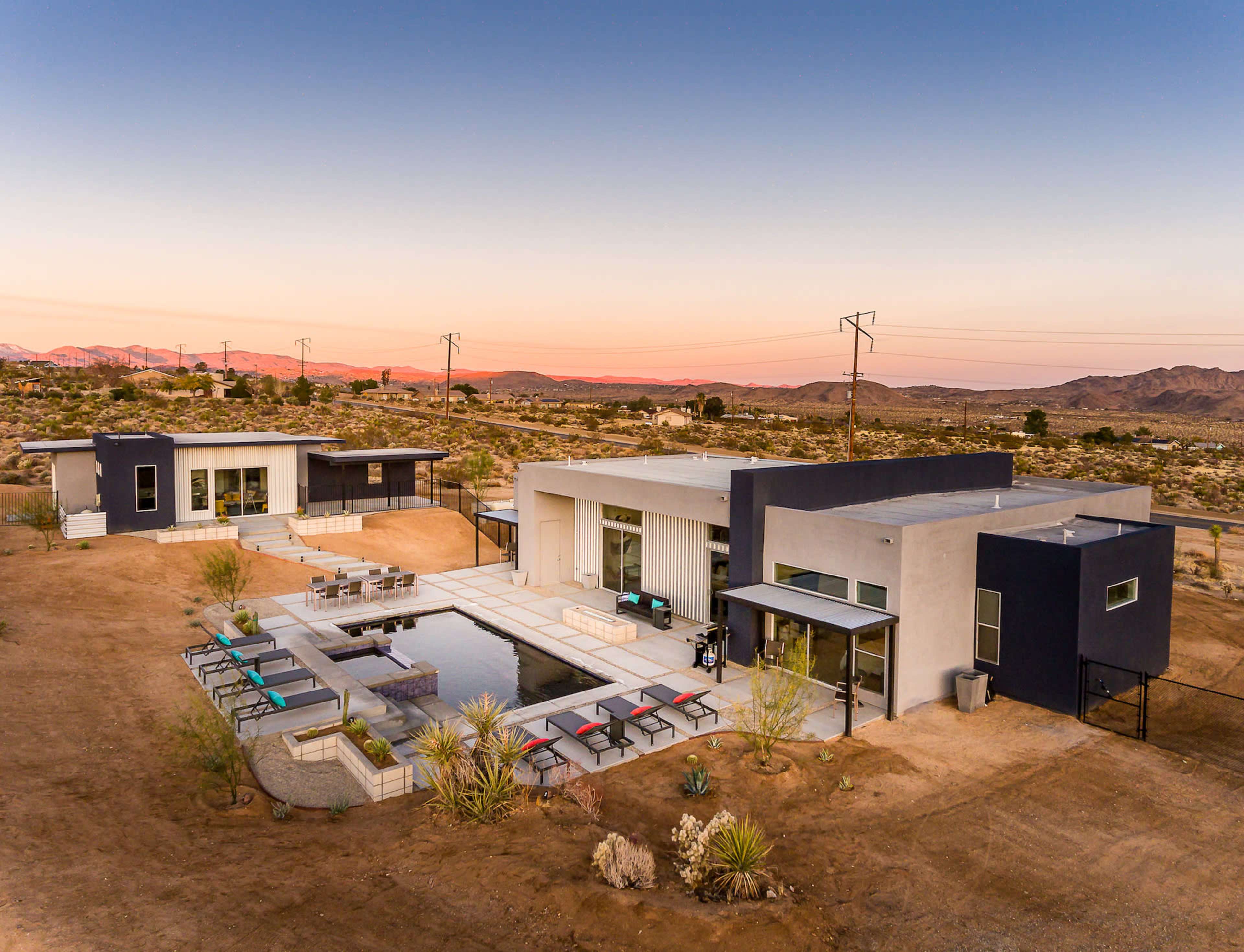 The image shows a modern desert residence with two buildings surrounding a central pool, set against a mountainous landscape during sunset.