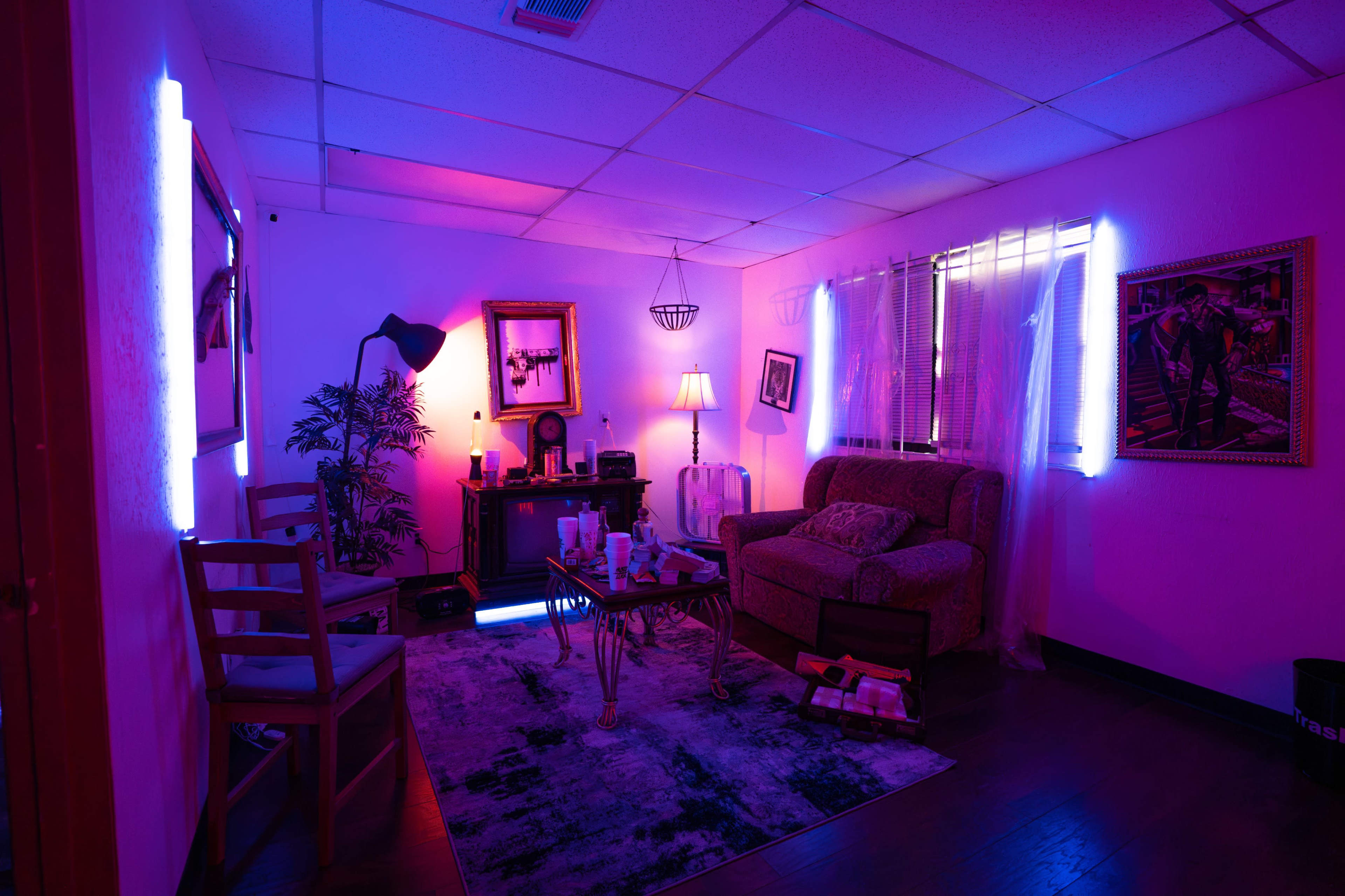 The image shows a dimly lit room with purple and blue lighting, featuring a couch, chairs, a coffee table, and decorative artwork on the walls.