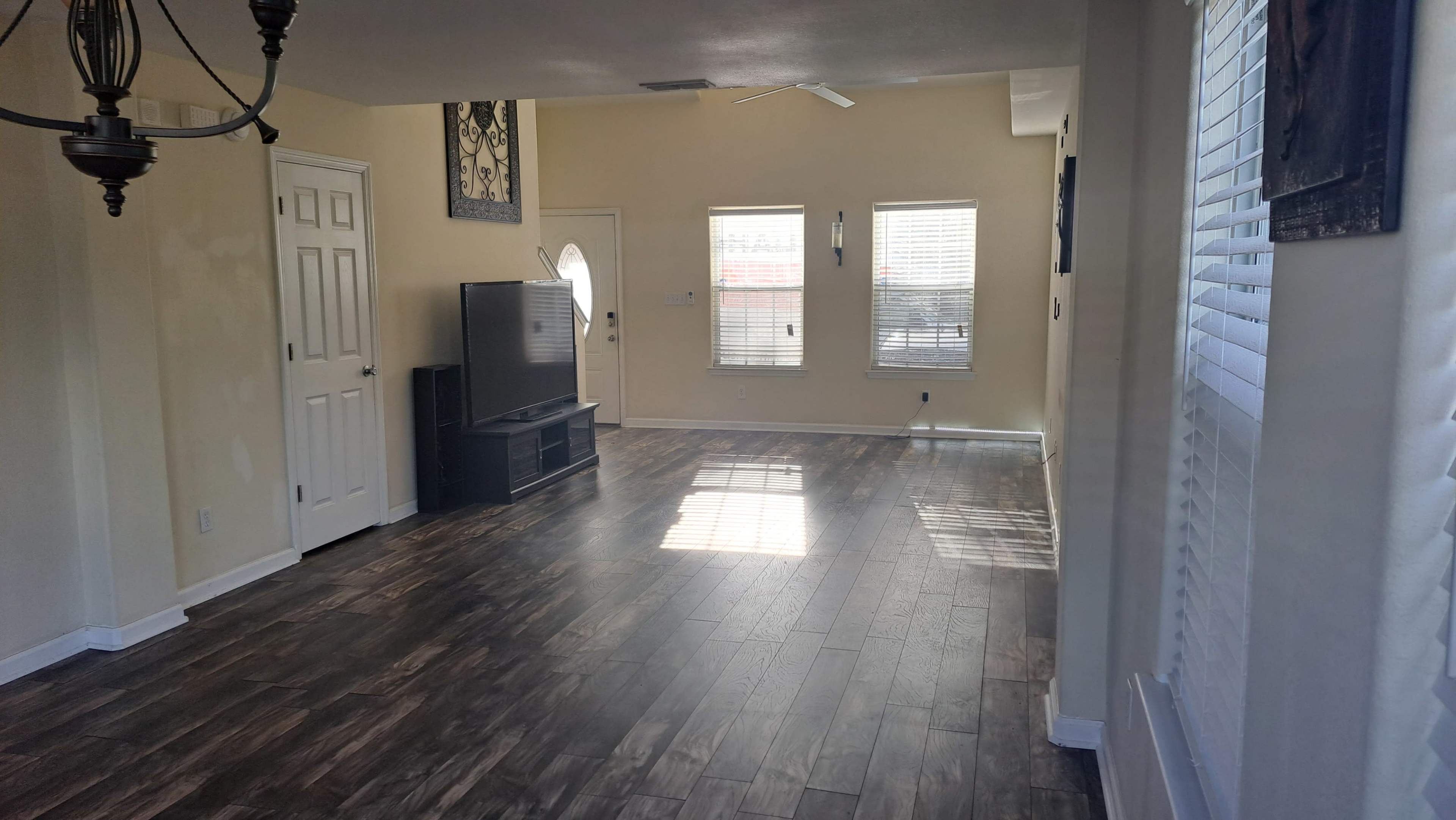 A spacious living room with wooden flooring, a large television, and multiple windows allowing natural light to enter.