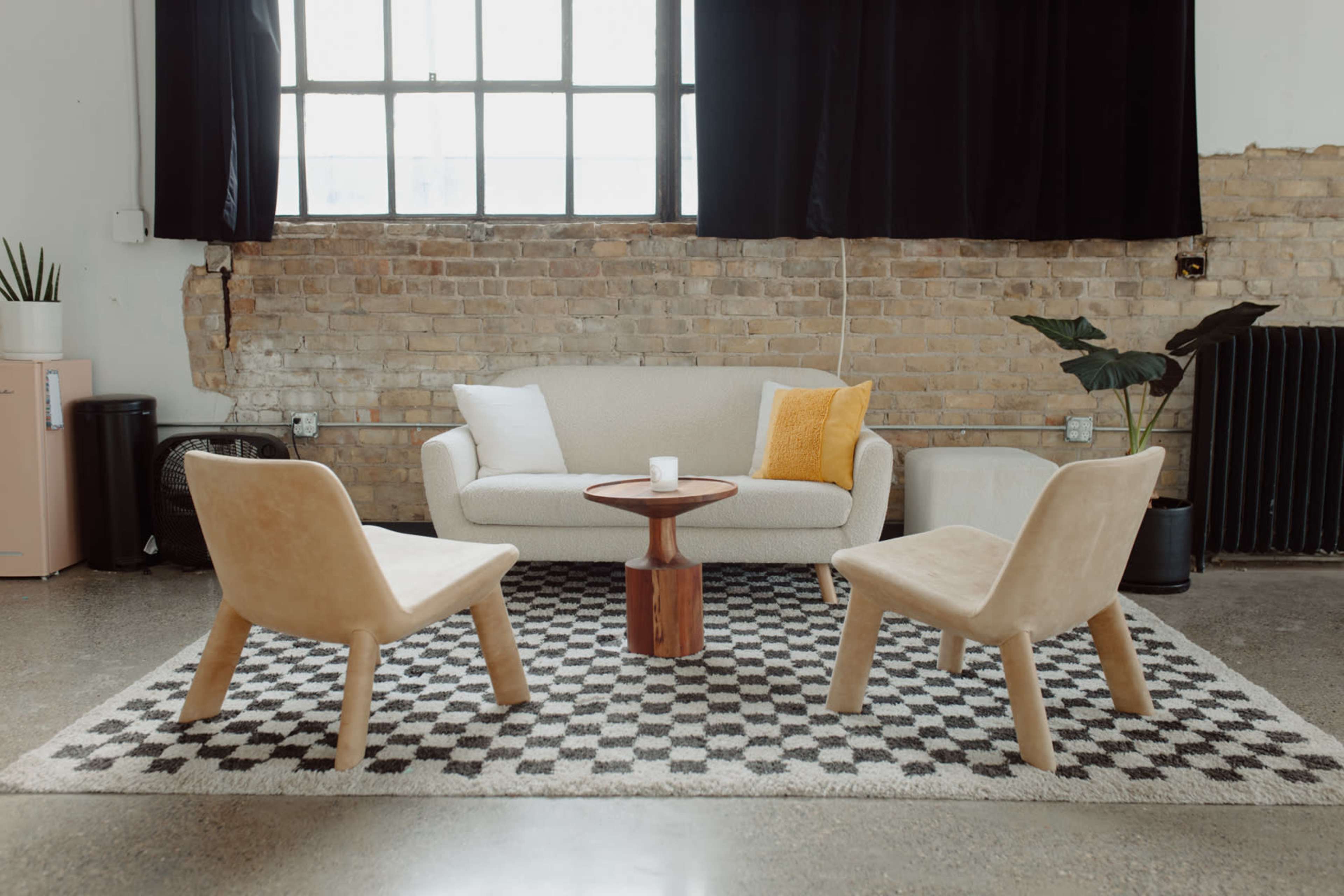A modern living space features a light-colored sofa, two beige chairs, a round wooden table, and a patterned rug against a backdrop of exposed brick walls and large windows.