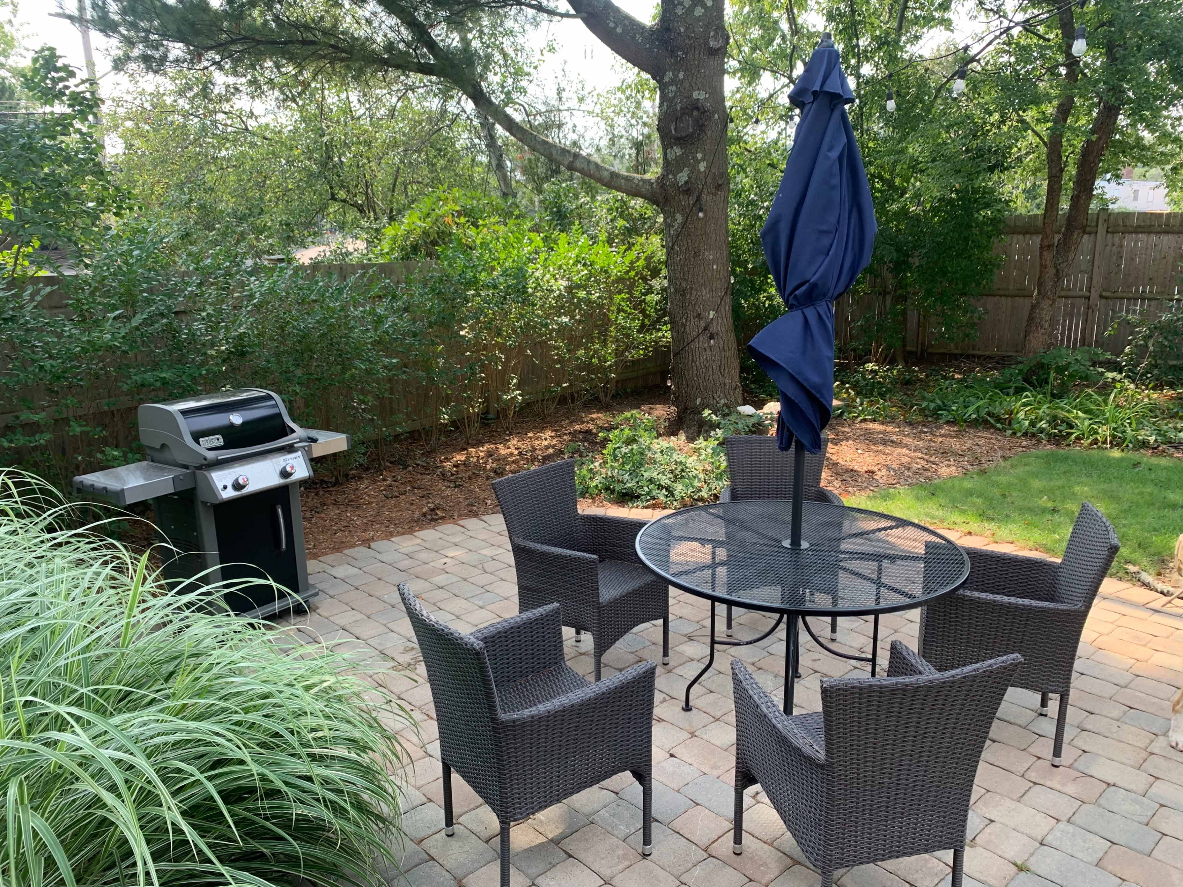A patio featuring a table with six chairs under an umbrella, alongside a grill and surrounded by lush greenery.