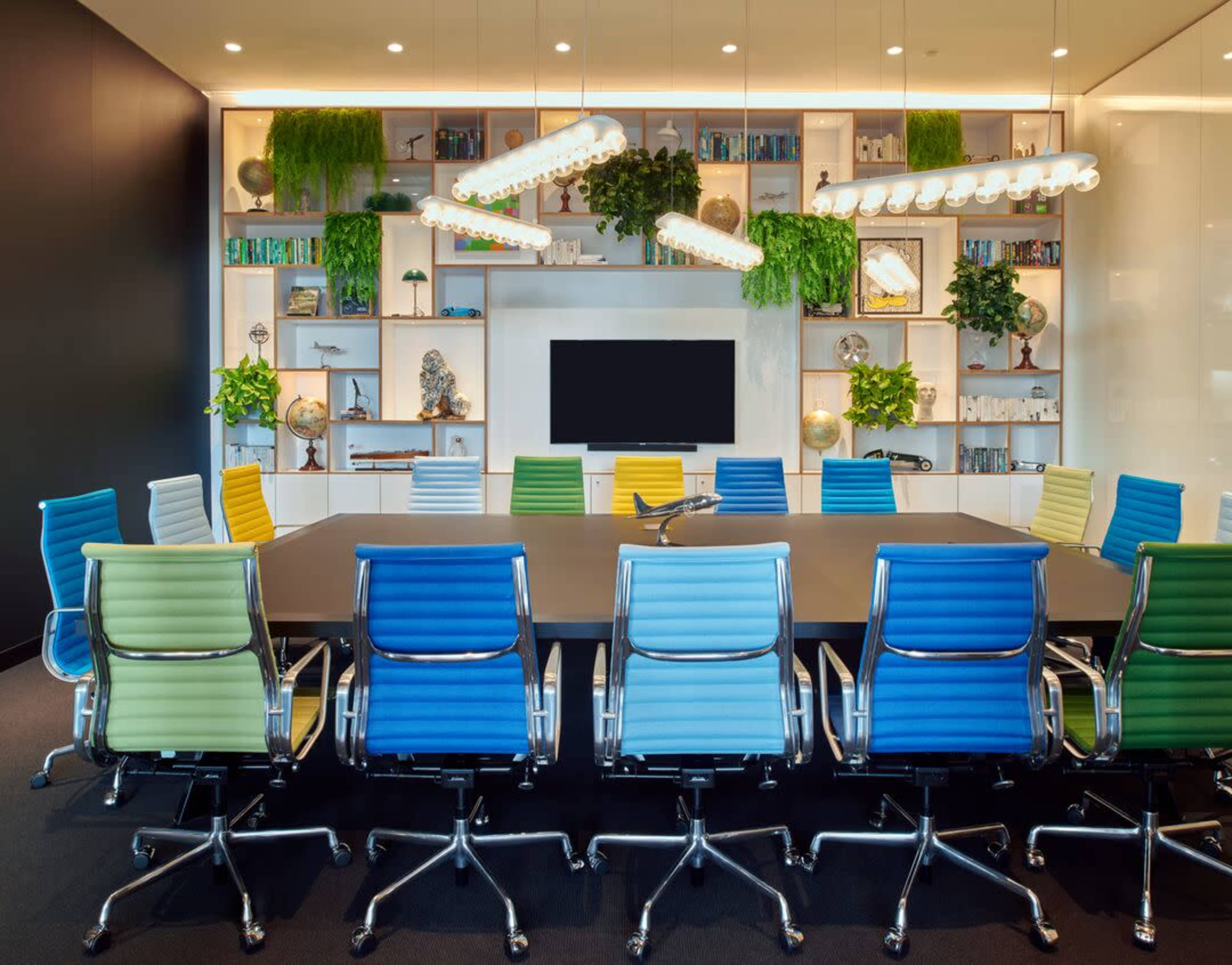 A modern conference room features a large table surrounded by colorful office chairs and a wall with shelves displaying plants and decorative items.