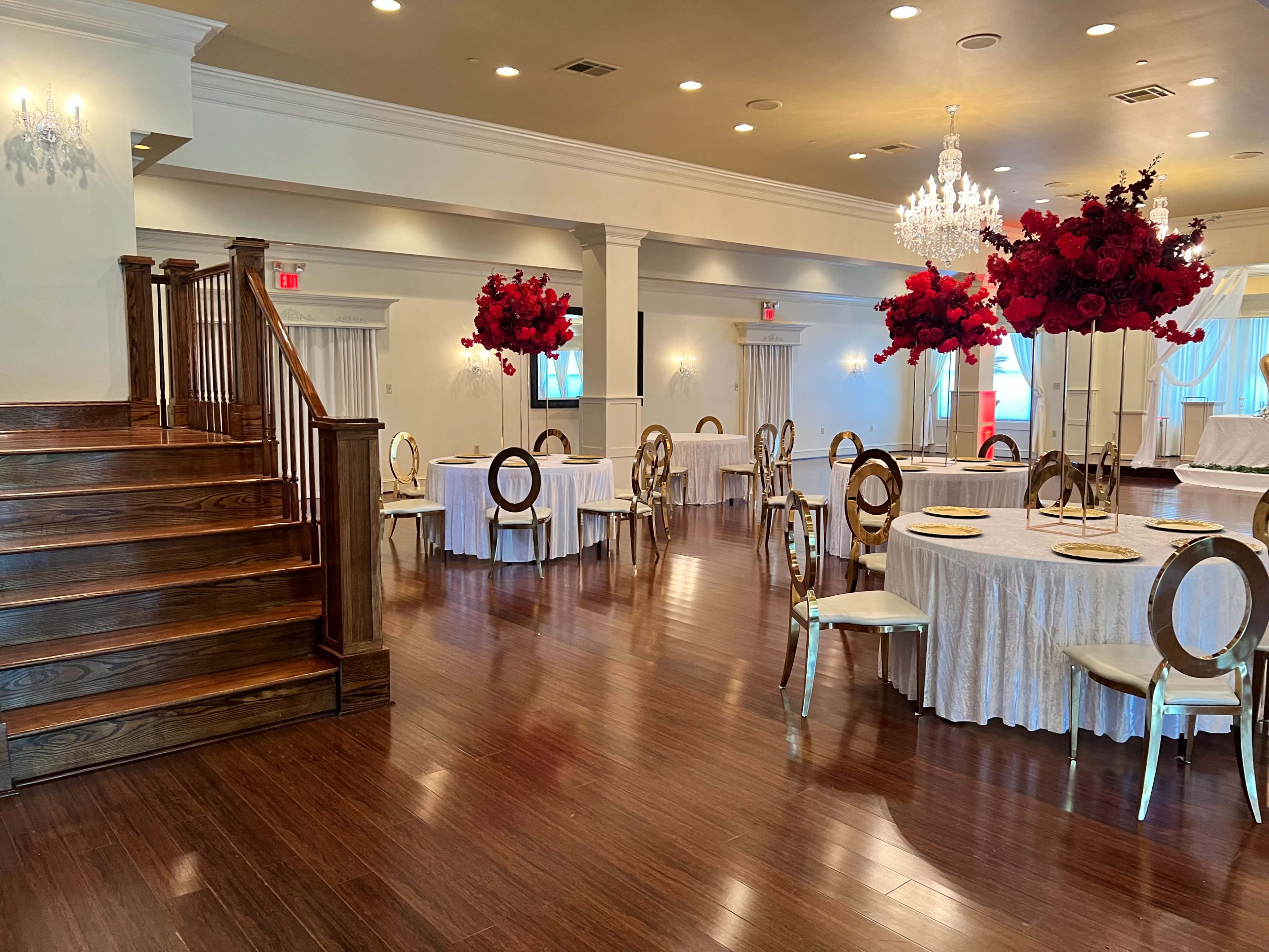 The image shows a spacious banquet hall with wooden flooring, round tables covered in white tablecloths, and tall floral centerpieces consisting of red roses.