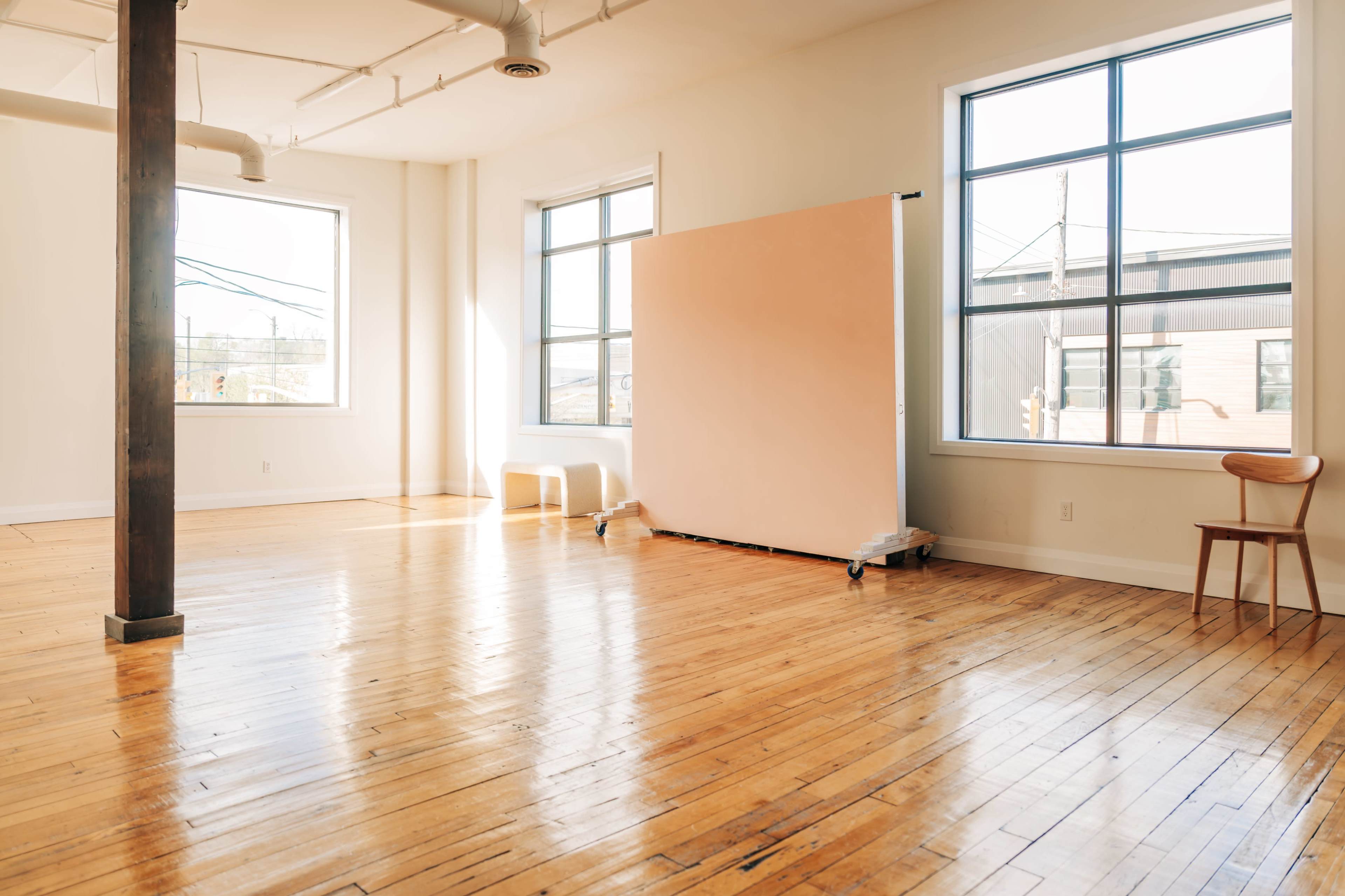 A bright, empty room features large windows, polished wooden floors, and a movable partition at one end.