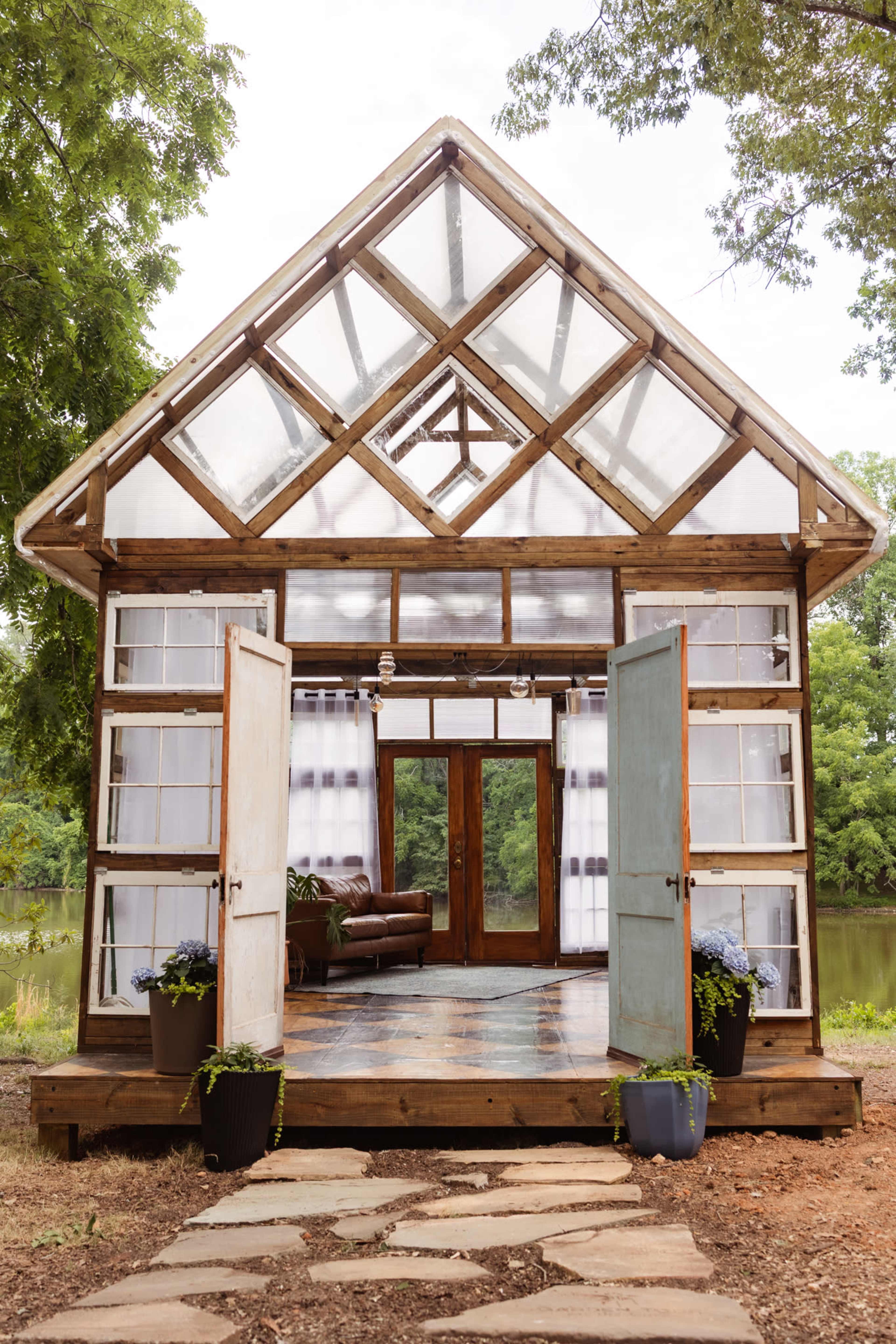 The image shows a glass-paneled cabin with a wooden frame, featuring open doors, a stone pathway, and surrounded by greenery.
