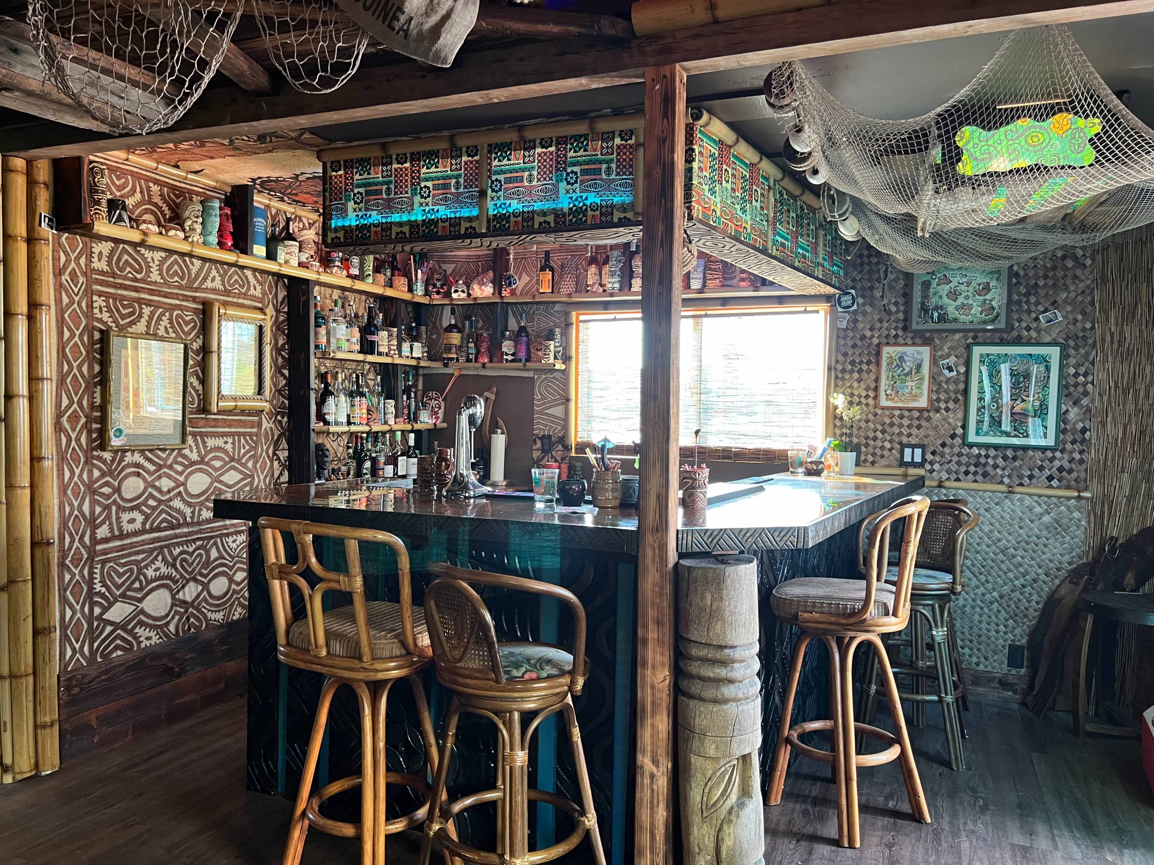 A tiki-themed bar with a bamboo frame, adorned shelves of colorful bottles, and a patterned wall design.