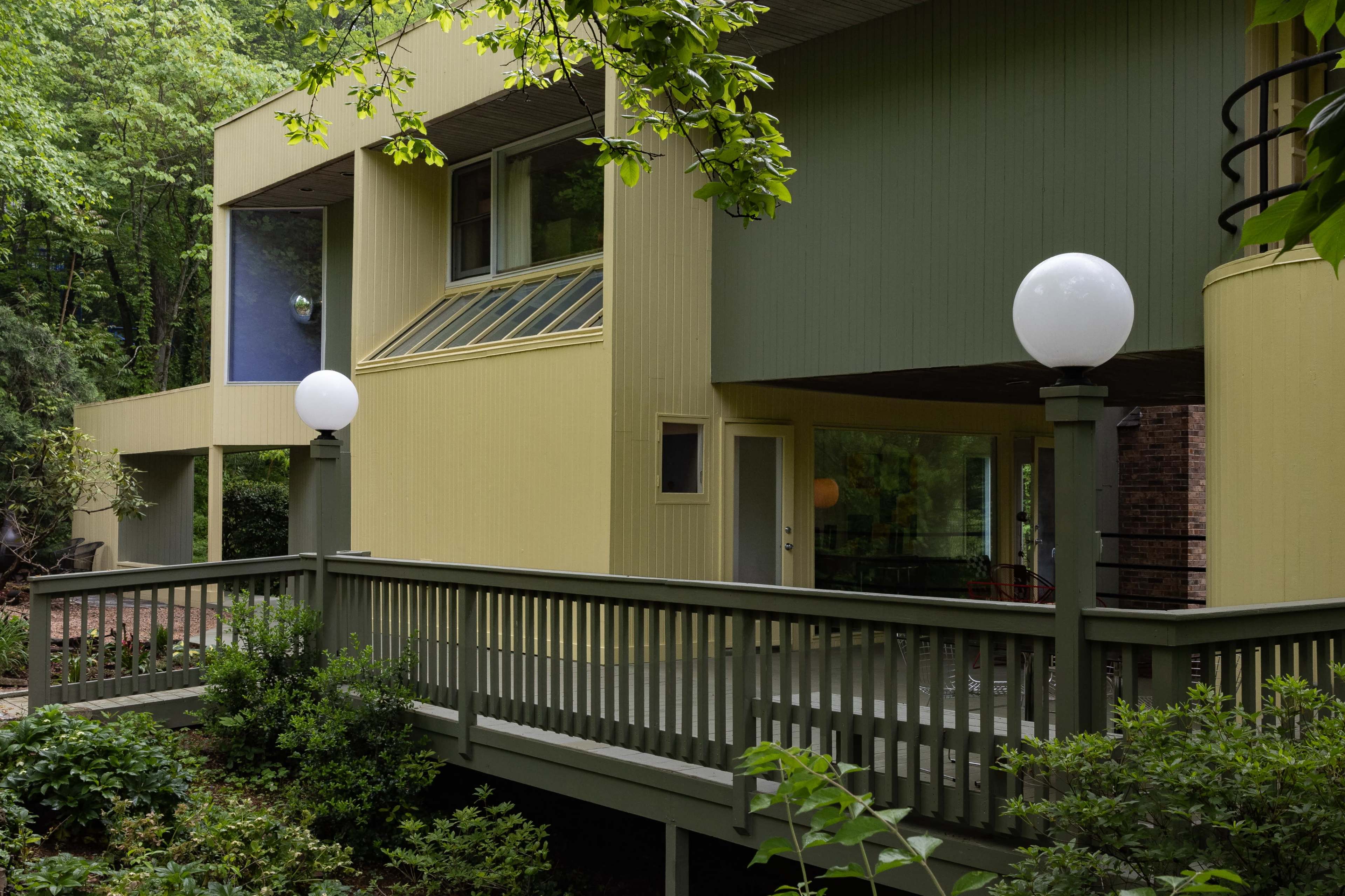 A modern, two-story house with a yellow and green exterior, surrounded by lush greenery and featuring round white lamp posts along a wooden deck.