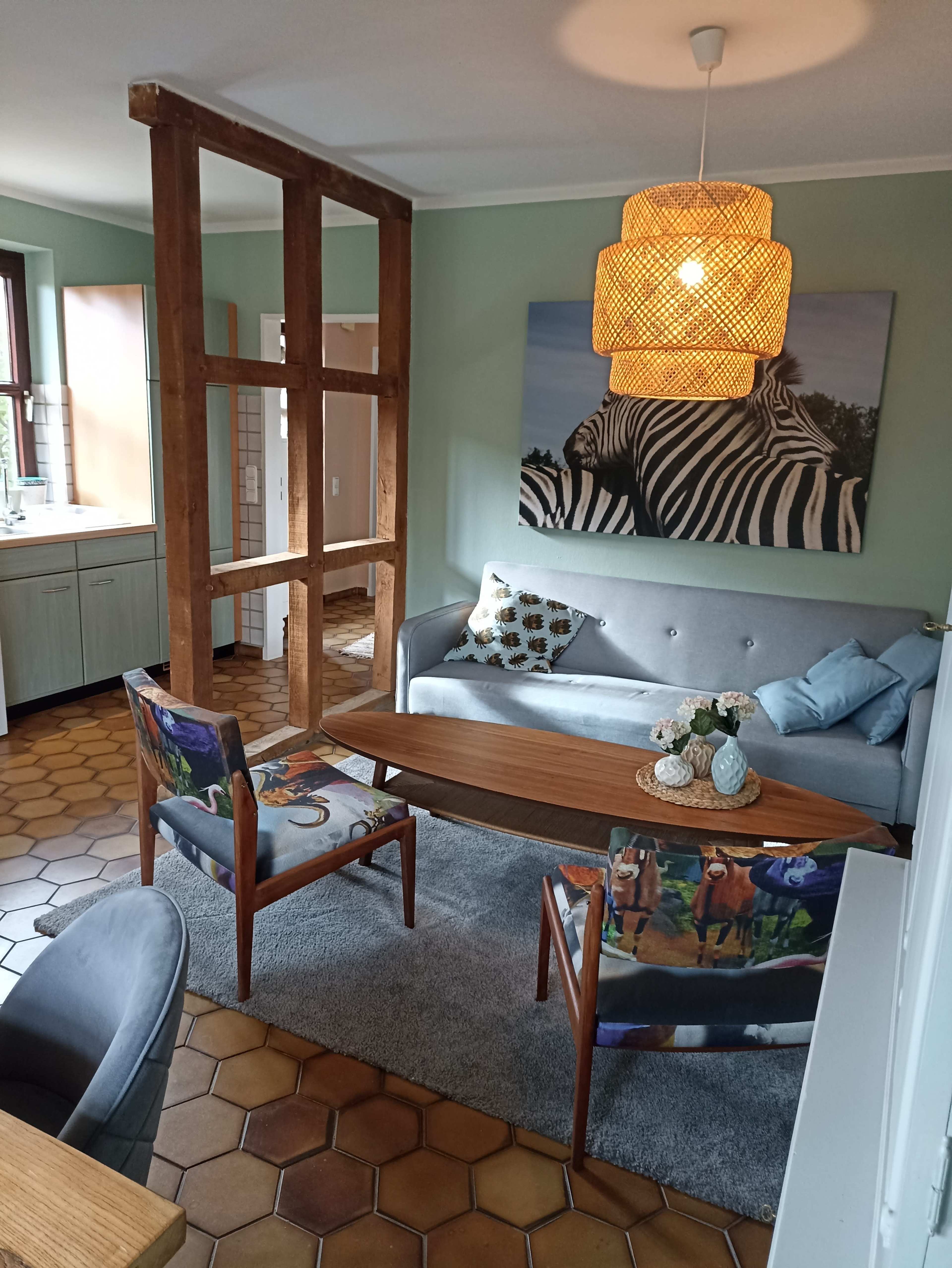 A living room features a gray sofa, a wooden divider, two patterned chairs, a wooden table with decorative items, and a large zebra print on the wall.