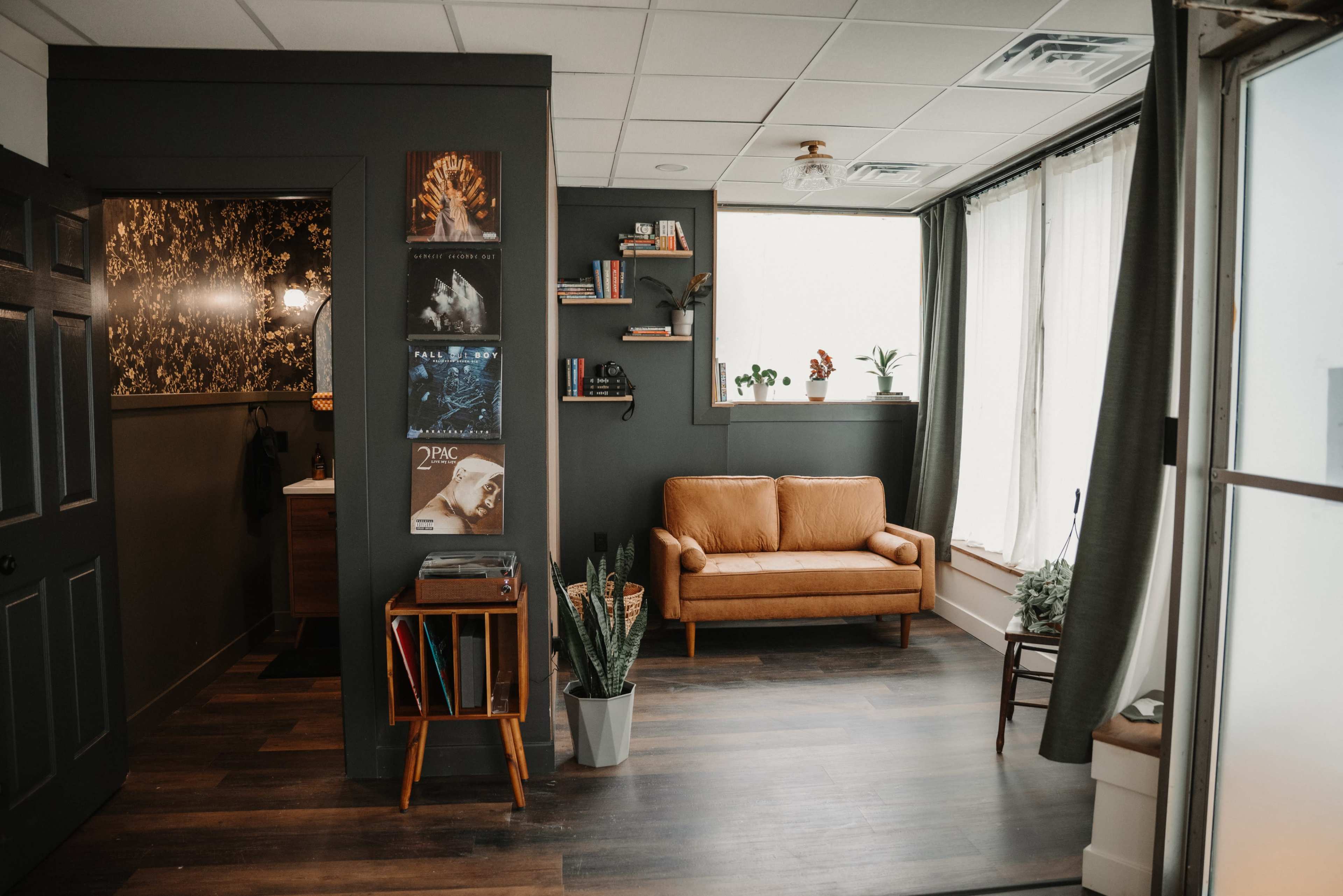 The image shows a cozy room with a brown couch, a bookshelf, and large windows allowing natural light to enter, while decorated with plants and artwork on the walls.