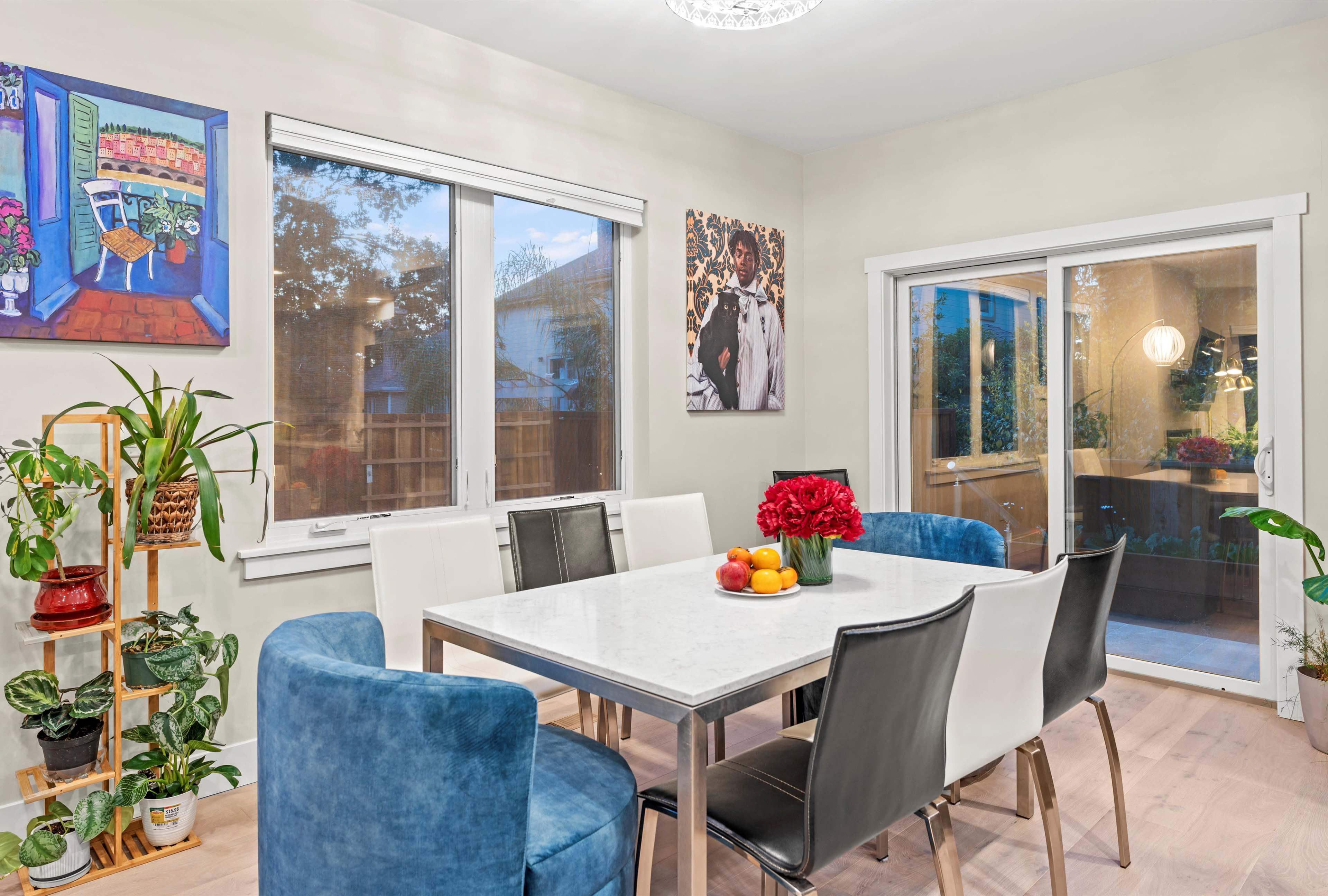A dining area features a white table surrounded by colorful chairs, with large windows letting in natural light and plants adding greenery.