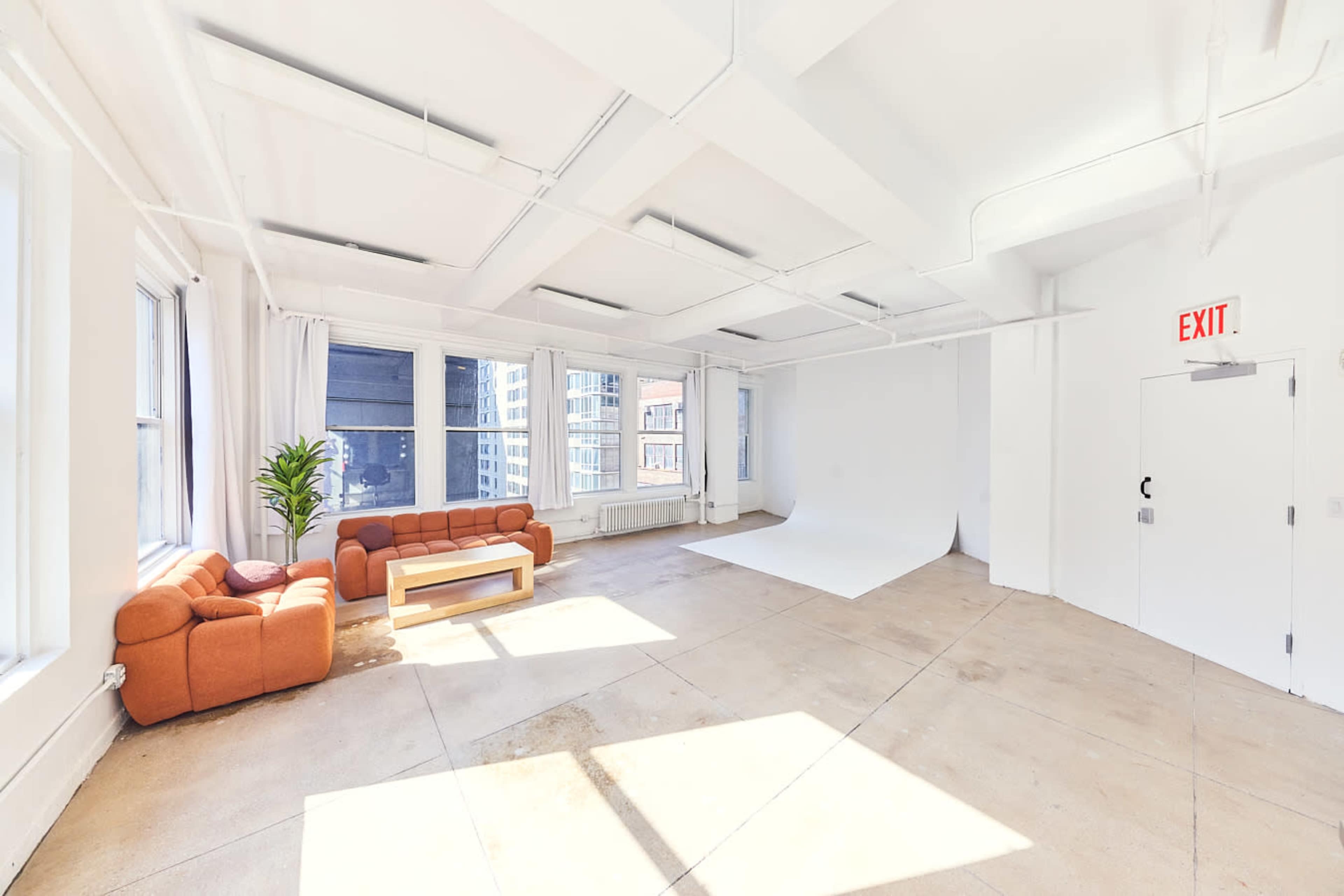 The image features a bright, spacious room with large windows, a brown couch and coffee table, and a white backdrop against a plain wall.