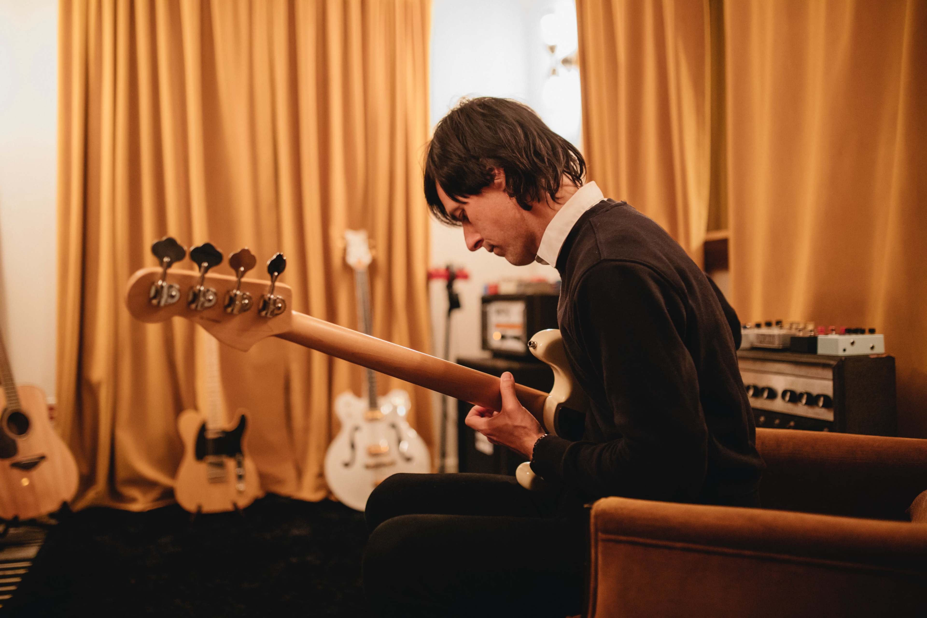 A musician sits on a chair in a room filled with various guitars, playing a bass guitar while surrounded by yellow curtains.