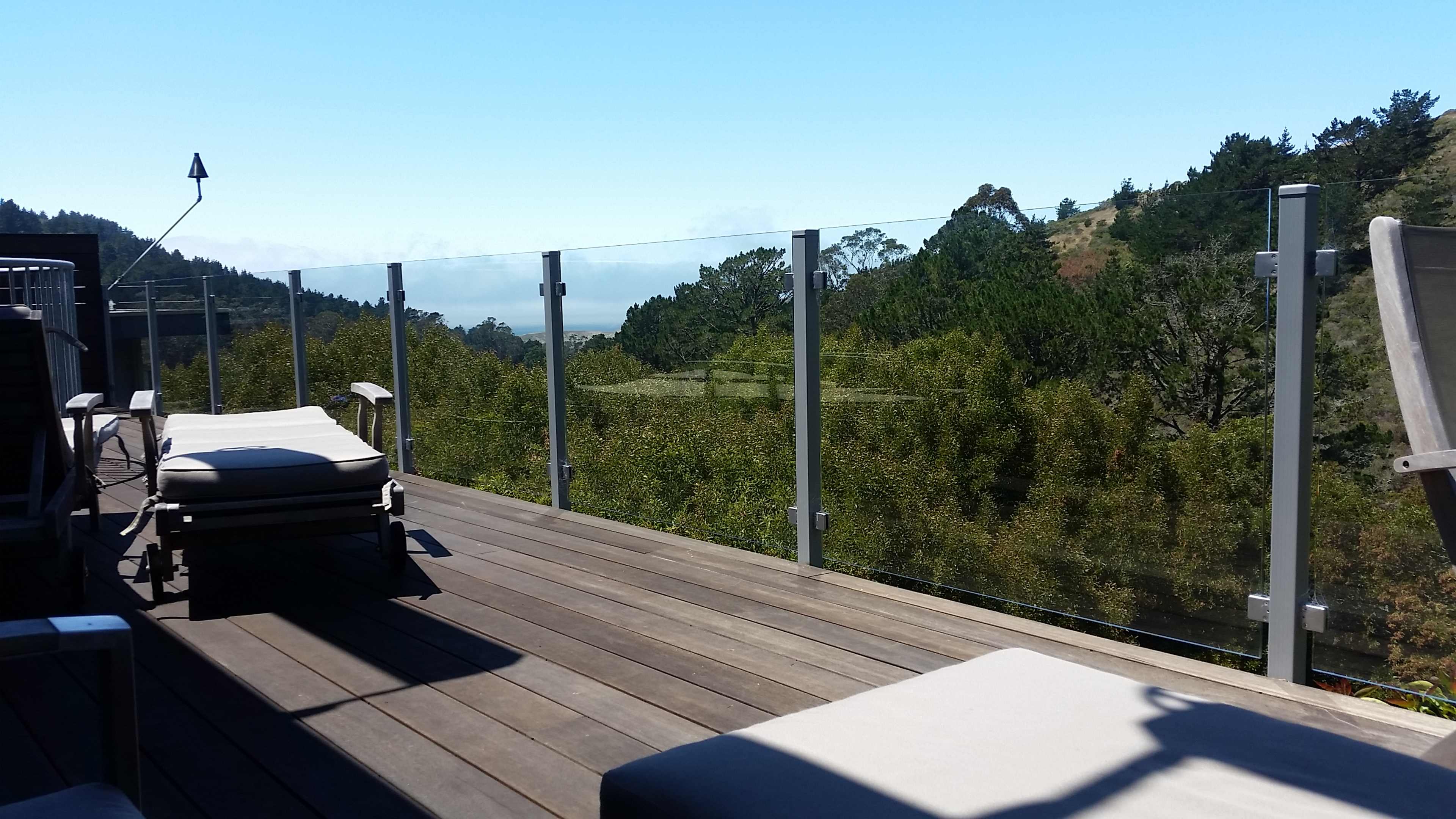 A wooden deck with large glass panels overlooks a green landscape and distant hills under a clear blue sky.