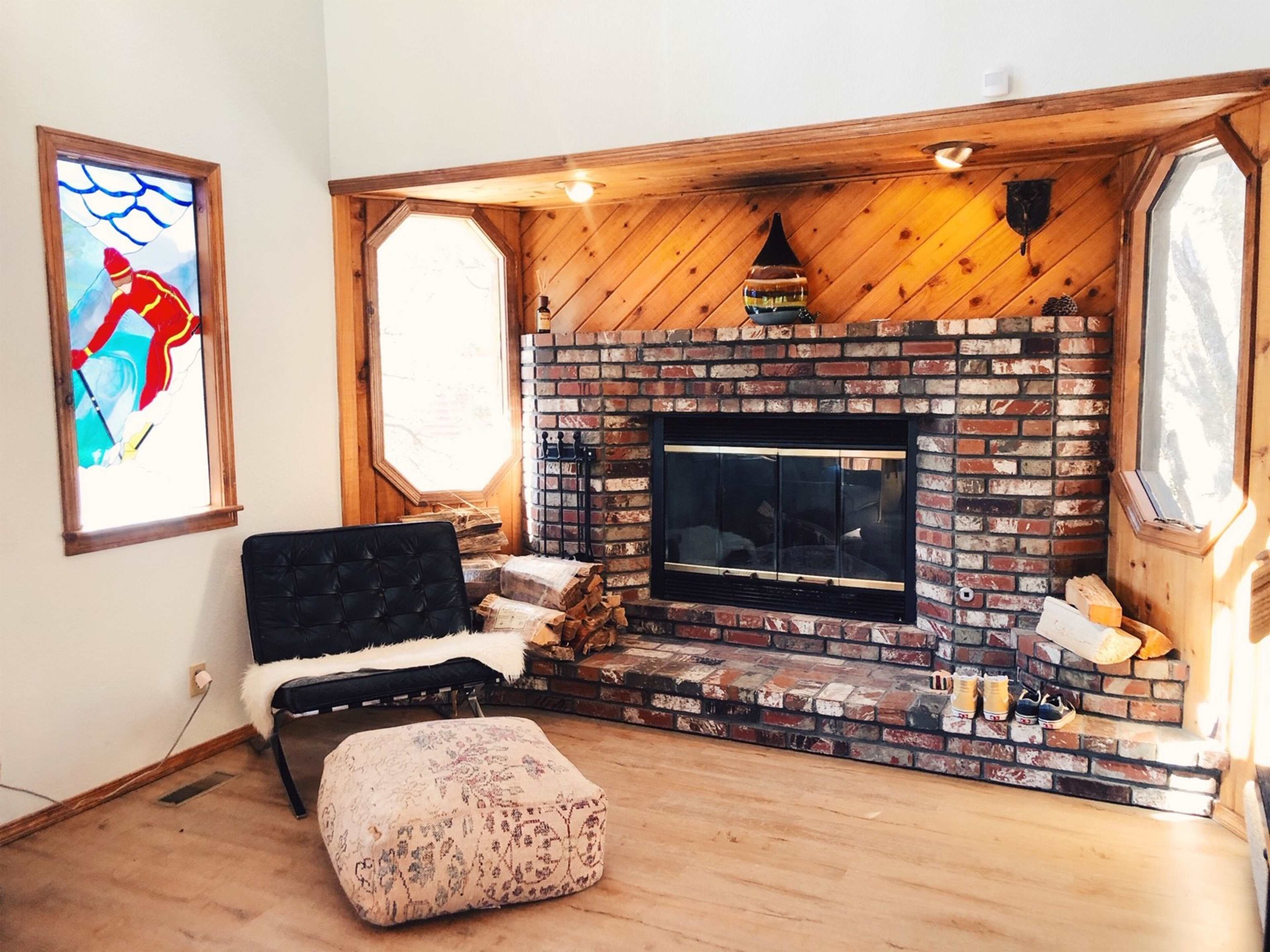 A brick fireplace is framed by wood paneling, with a quilted ottoman and a black chair nearby, illuminated by natural light from a stained glass window depicting a skier.