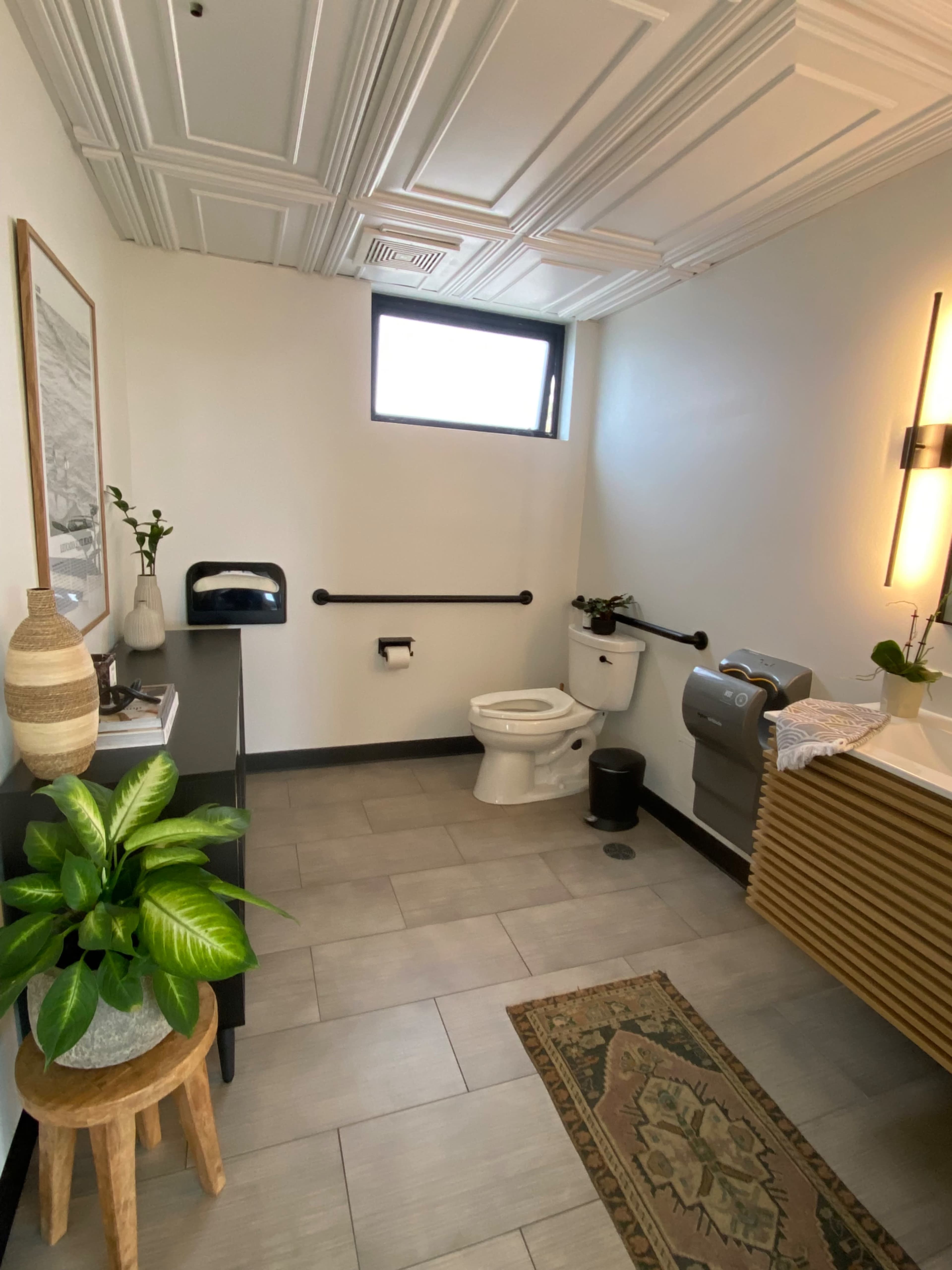 A small bathroom features a toilet, a washbasin with wooden storage, and a window, with a decorative plant and rug enhancing the space.
