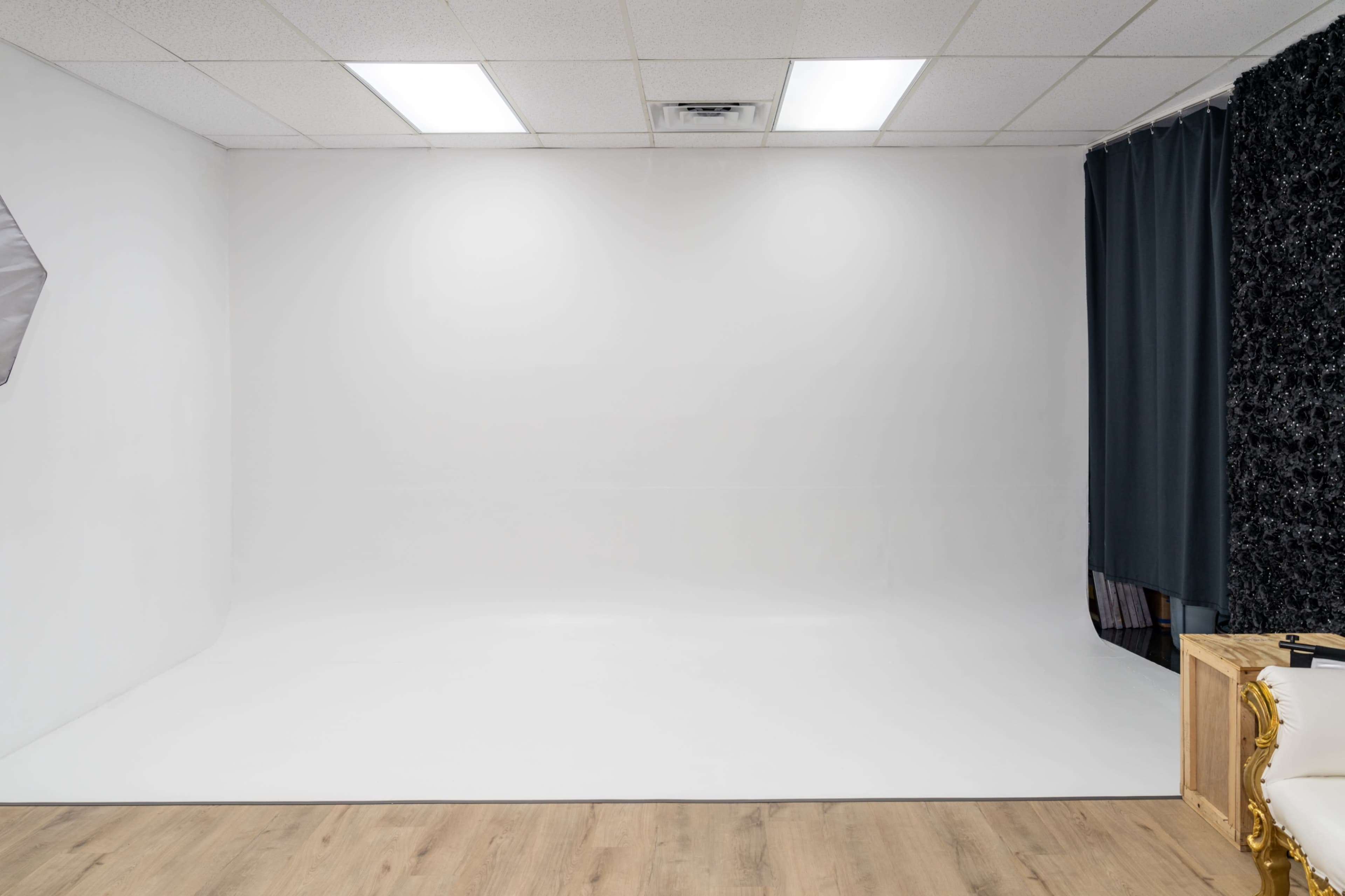 The image shows an empty studio space with a white backdrop, wooden flooring, and a black curtain on one side.