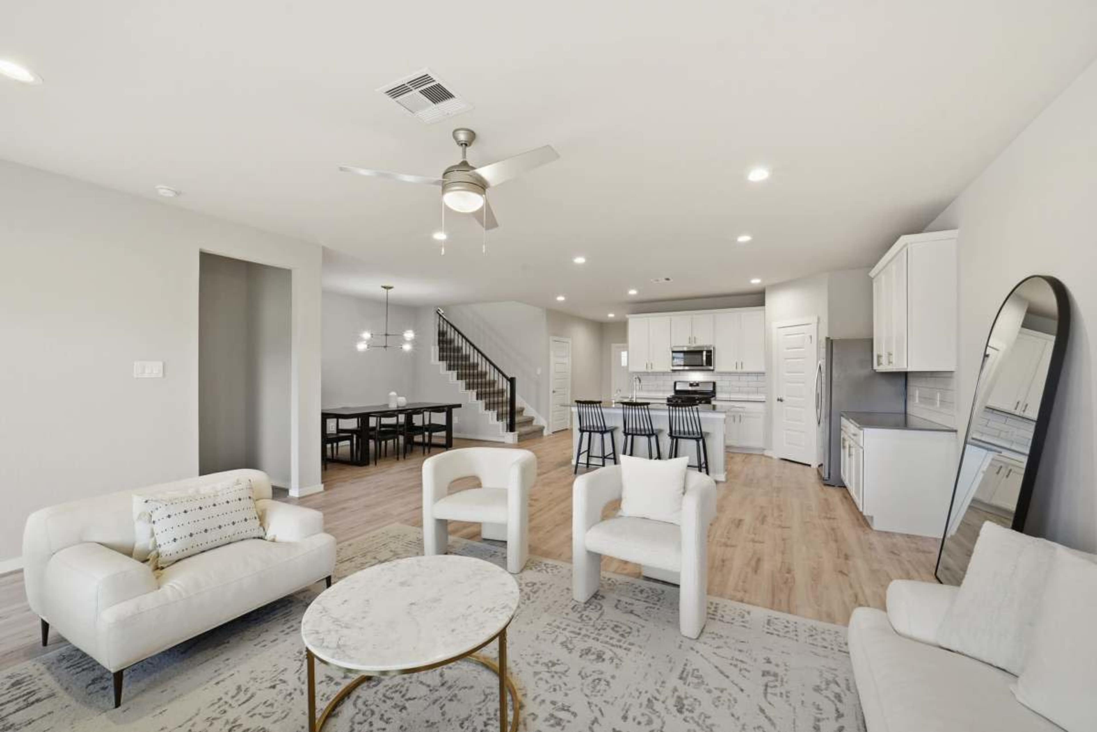 The image shows a spacious, modern living room connected to a dining area and kitchen, featuring light-colored furniture and a staircase in the background.
