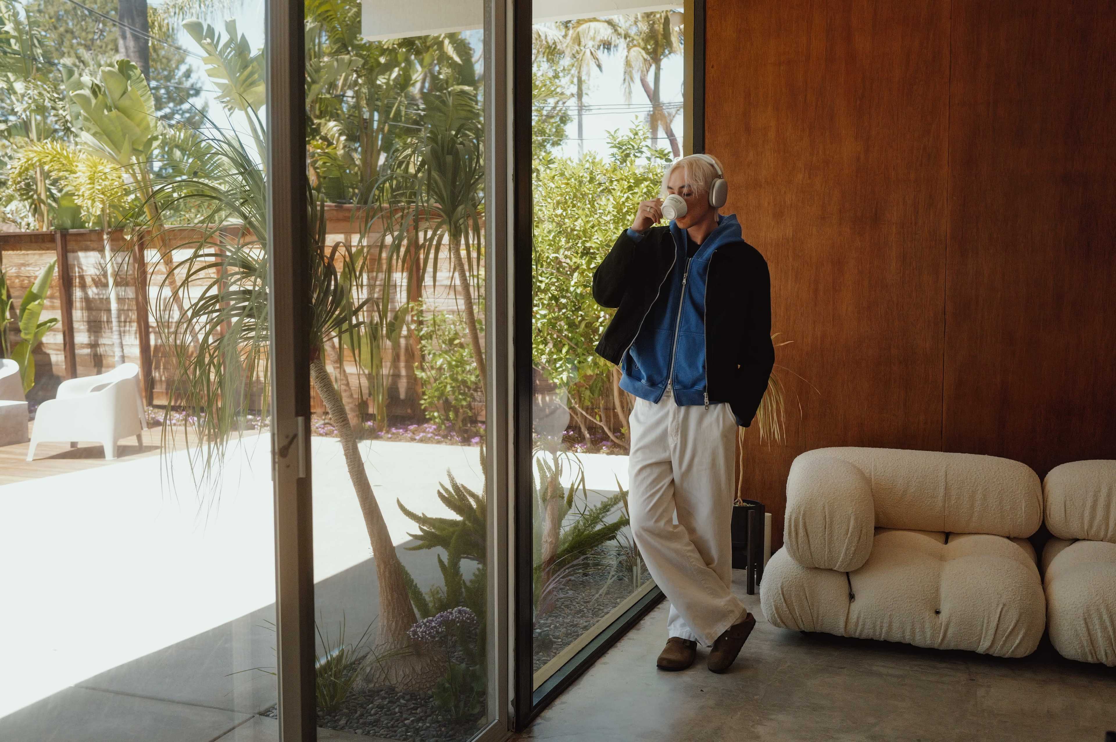 A person wearing headphones stands by a large glass window in a modern room, talking on the phone while surrounded by indoor plants.