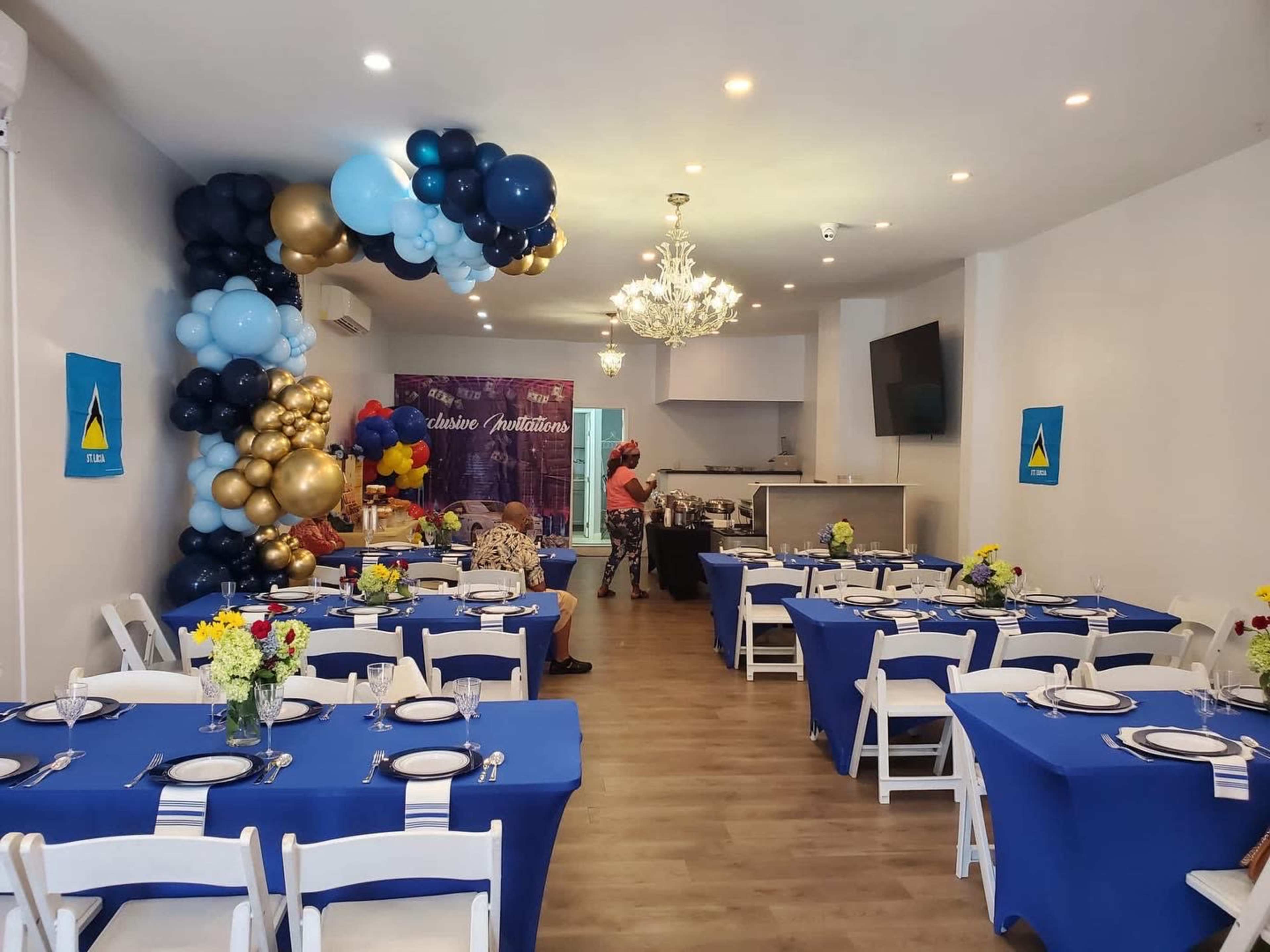 A brightly decorated indoor venue features tables with blue tablecloths set for a celebration, adorned with floral centerpieces and a balloon arch in the background.