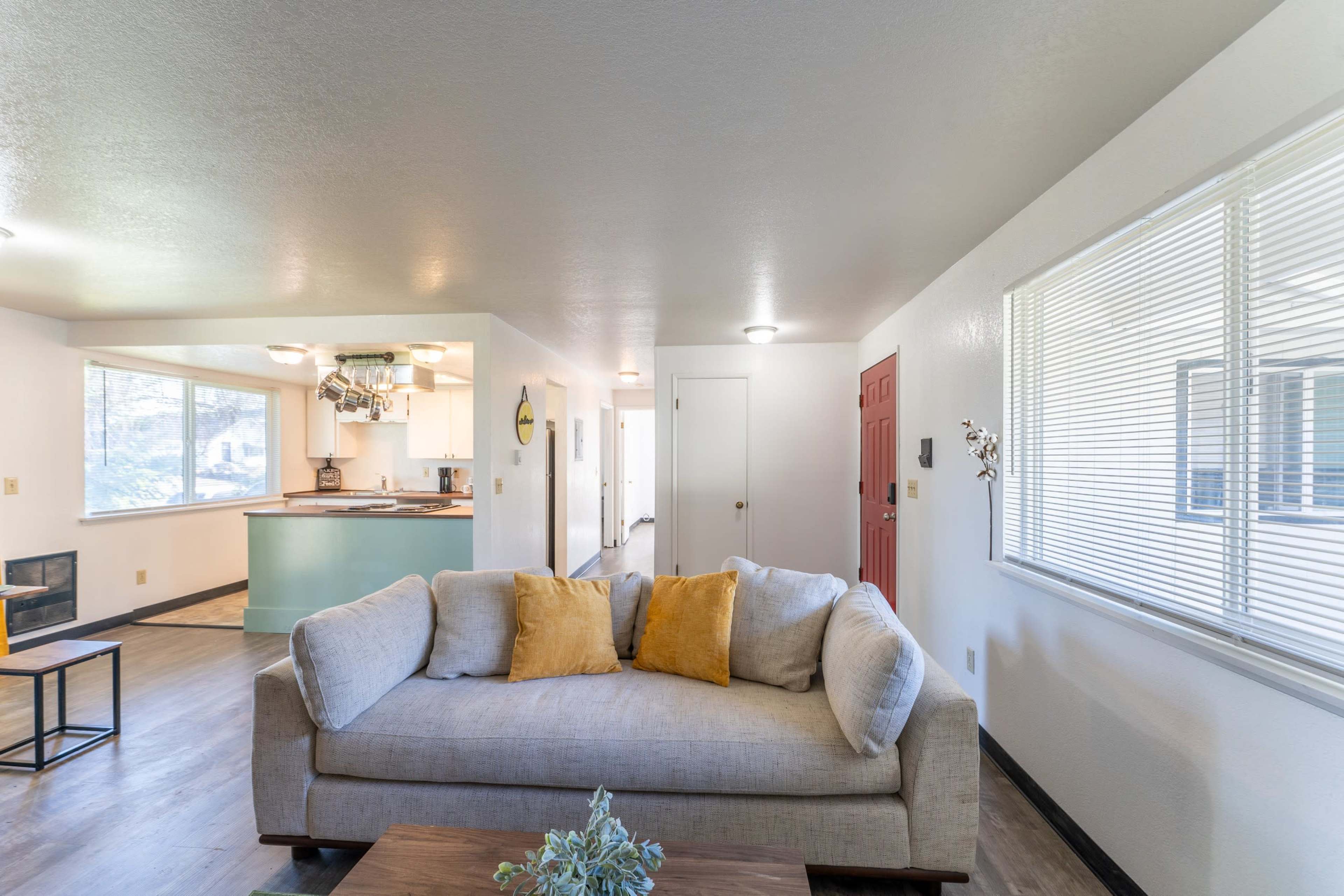 A living room features a light-colored sofa with yellow throw pillows, facing a wooden coffee table, and an open kitchen is visible in the background.