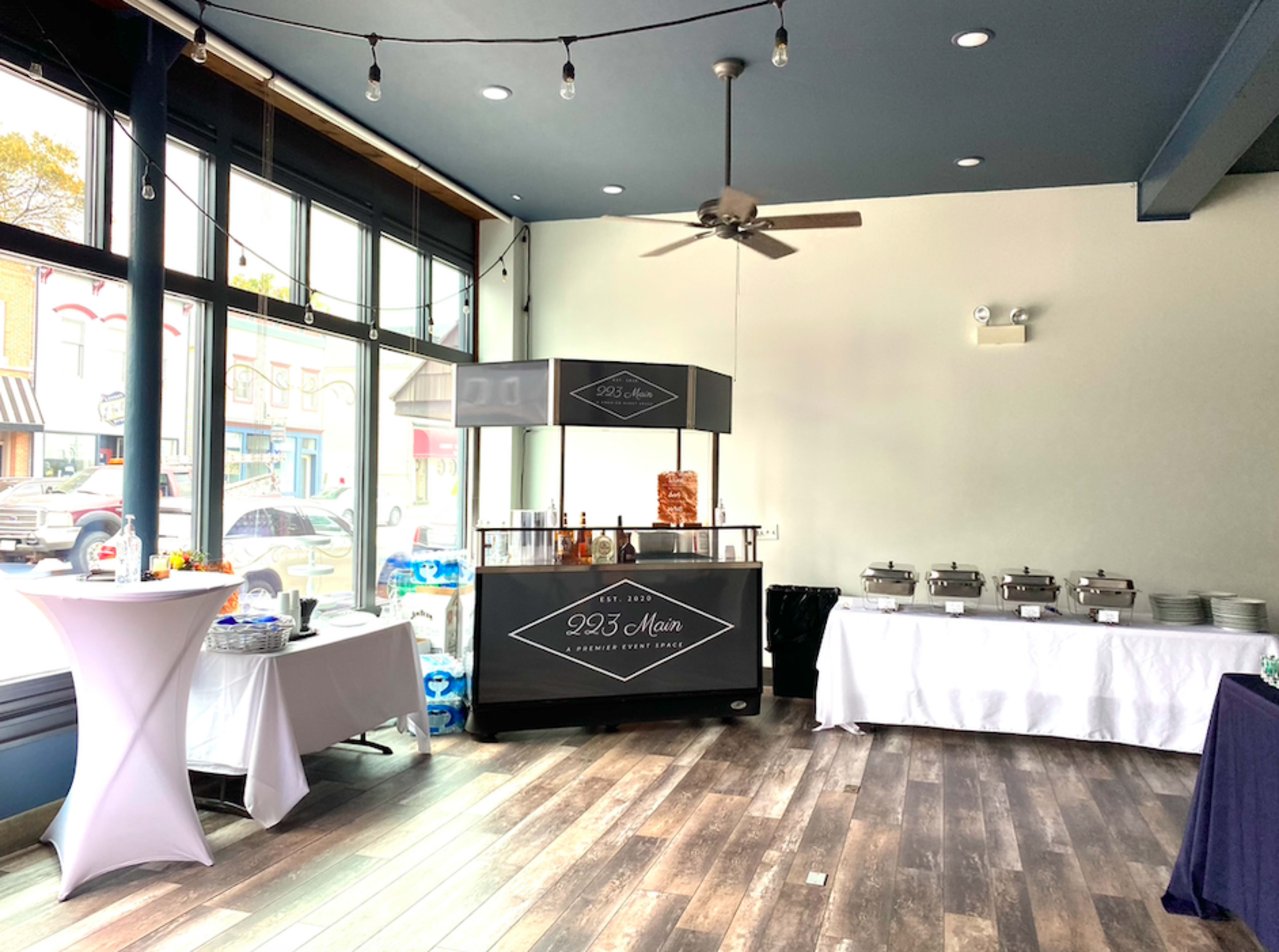The image shows a spacious event venue featuring a food setup with two catering tables, a portable bar with a sign that reads "923 Main," and large windows allowing natural light to enter.