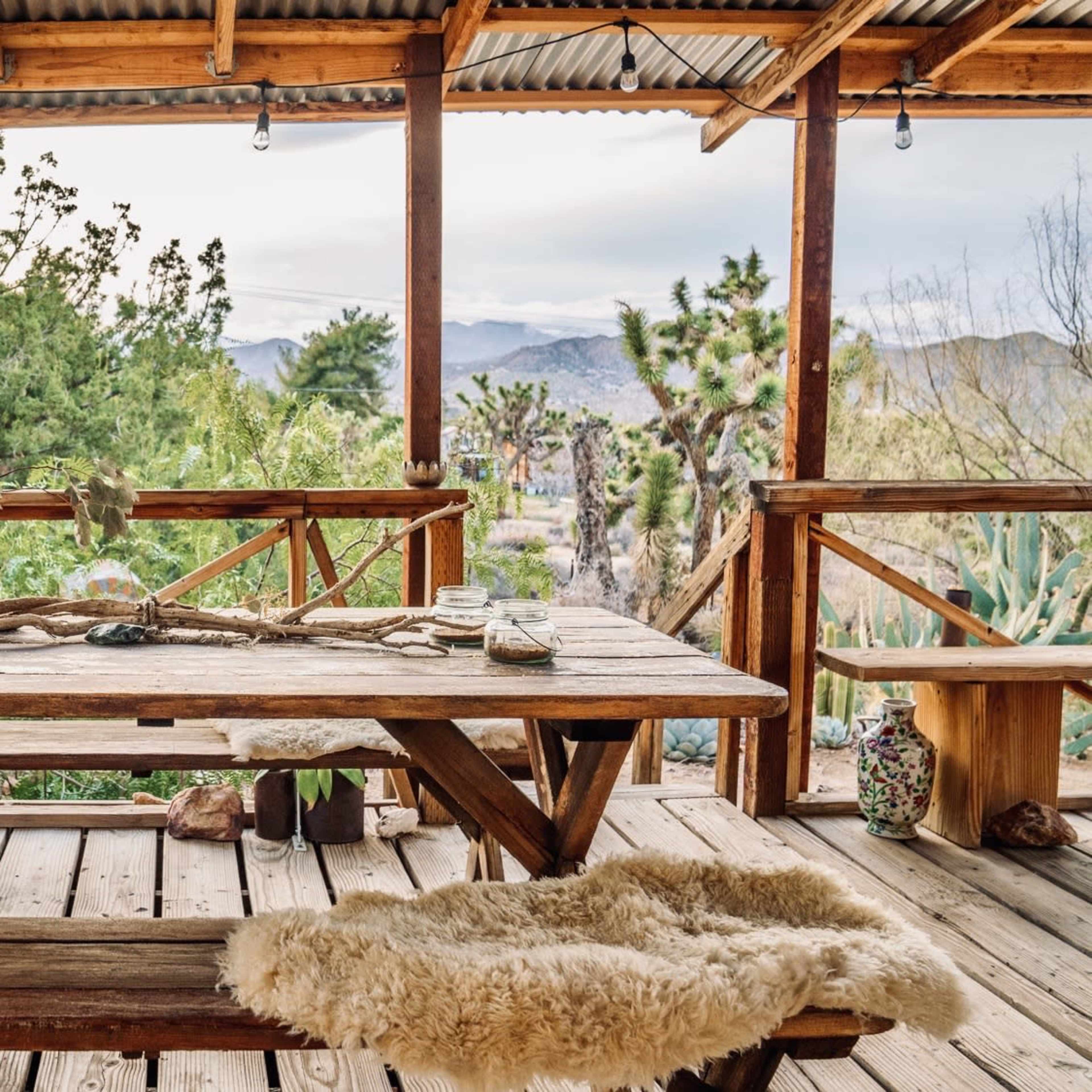 A wooden table and benches are set up on a covered porch overlooking a desert landscape with Joshua trees and mountains in the distance.